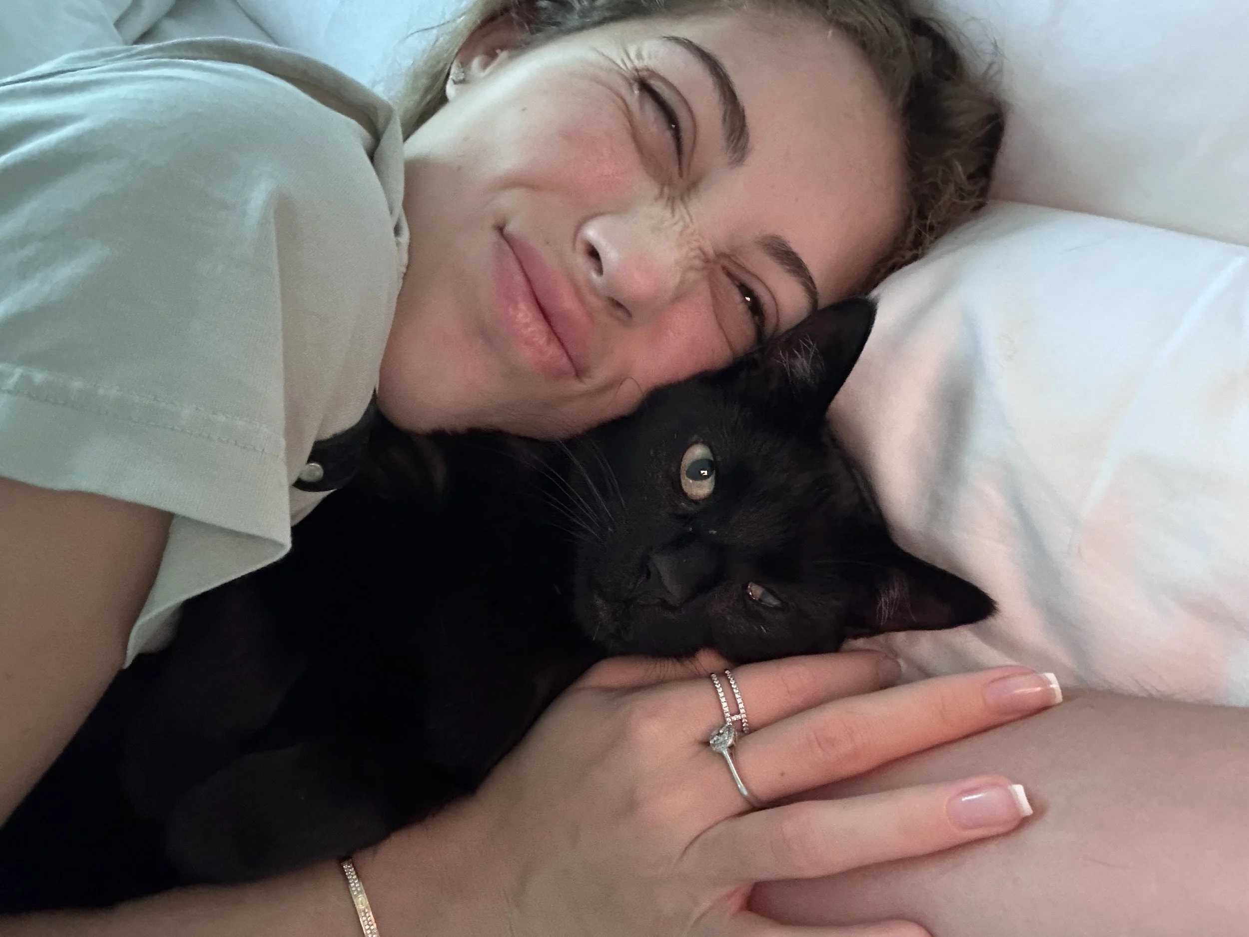A woman smiling and lying on a bed next to a black cat, with her arm around the cat.