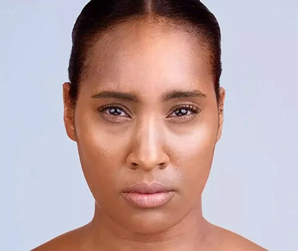 Close-up portrait of an African American woman with a serious expression, minimal makeup, against a light blue background.