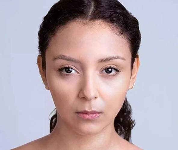 Close-up of a woman with brown hair pulled back, wearing subtle makeup, with a neutral expression against a light gray background.