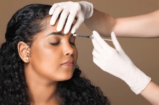 A woman with curly dark hair smiling as a gloved hand administers a cosmetic injection to her face.