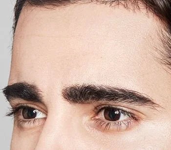 Close-up of a person's forehead and eyes, showing thick eyebrows and dark hair.