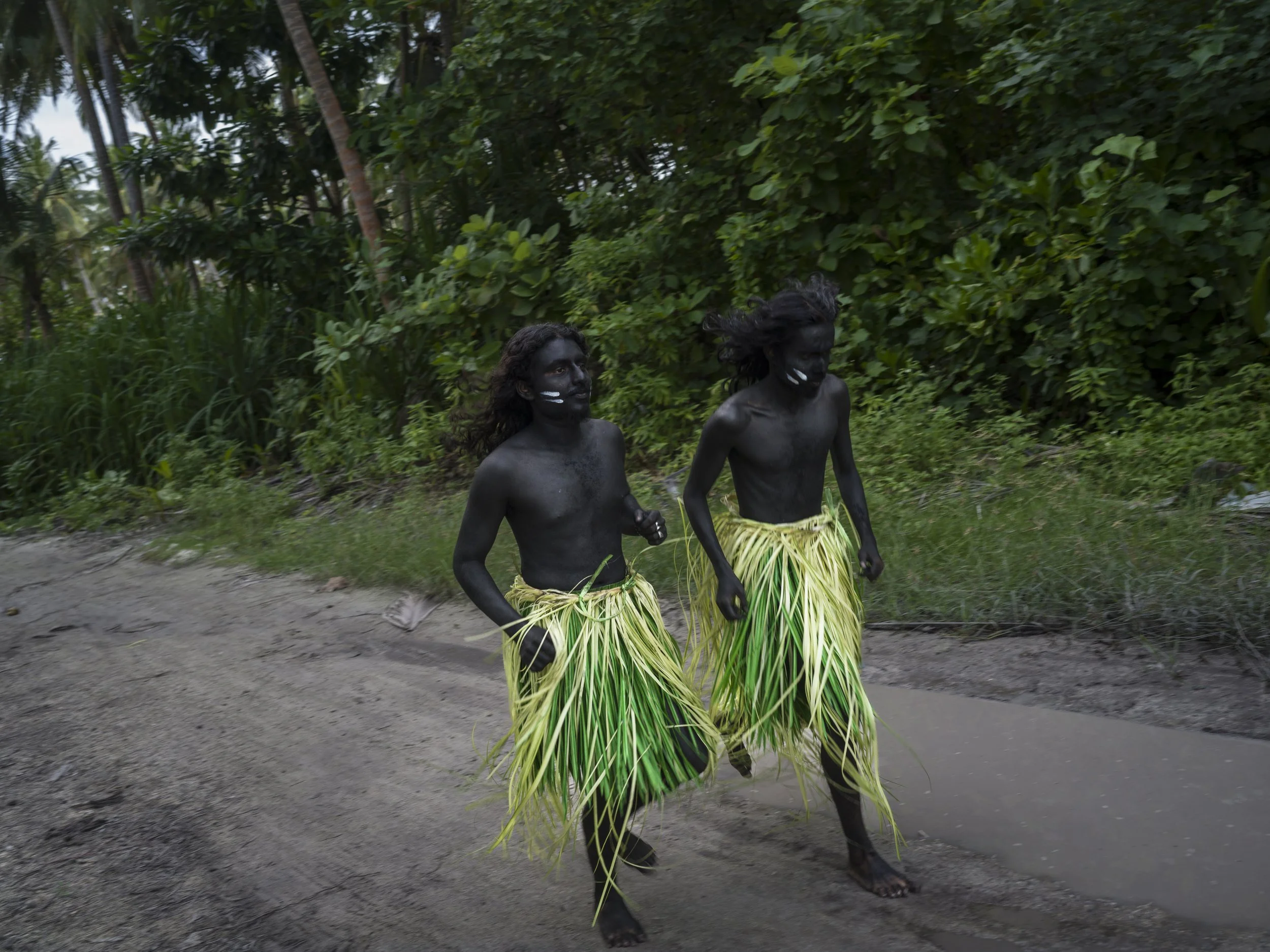 Two men with black painted bodies, wearing grass skirts and walking along a dirt path surrounded by lush green vegetation.