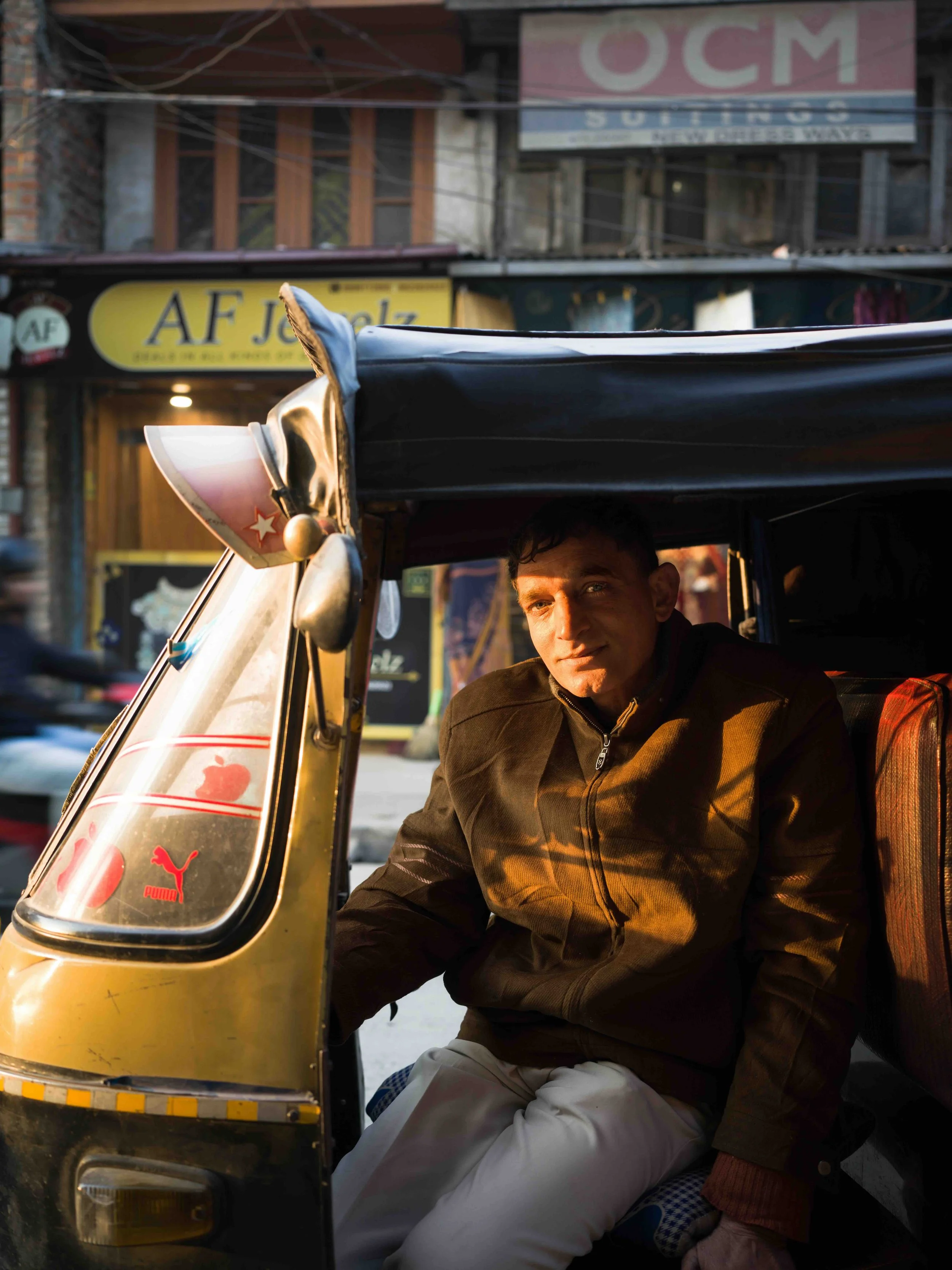 A Kashmiri man seated inside a yellow auto rickshaw at sunset in an urban street with shops and signs in the background, in Srinagar, Kashmir