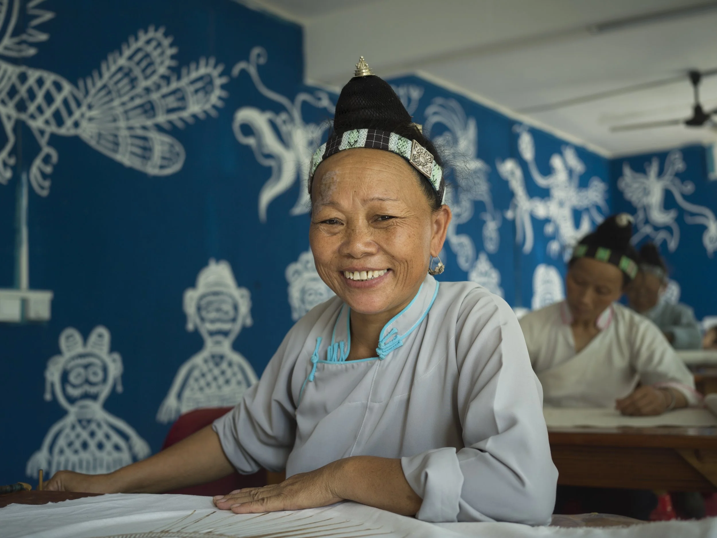 Smiling woman wearing traditional attire, sitting at a craft table. In the background, other women are engaged in similar activities, with a blue wall decorated with white cultural and artistic designs.