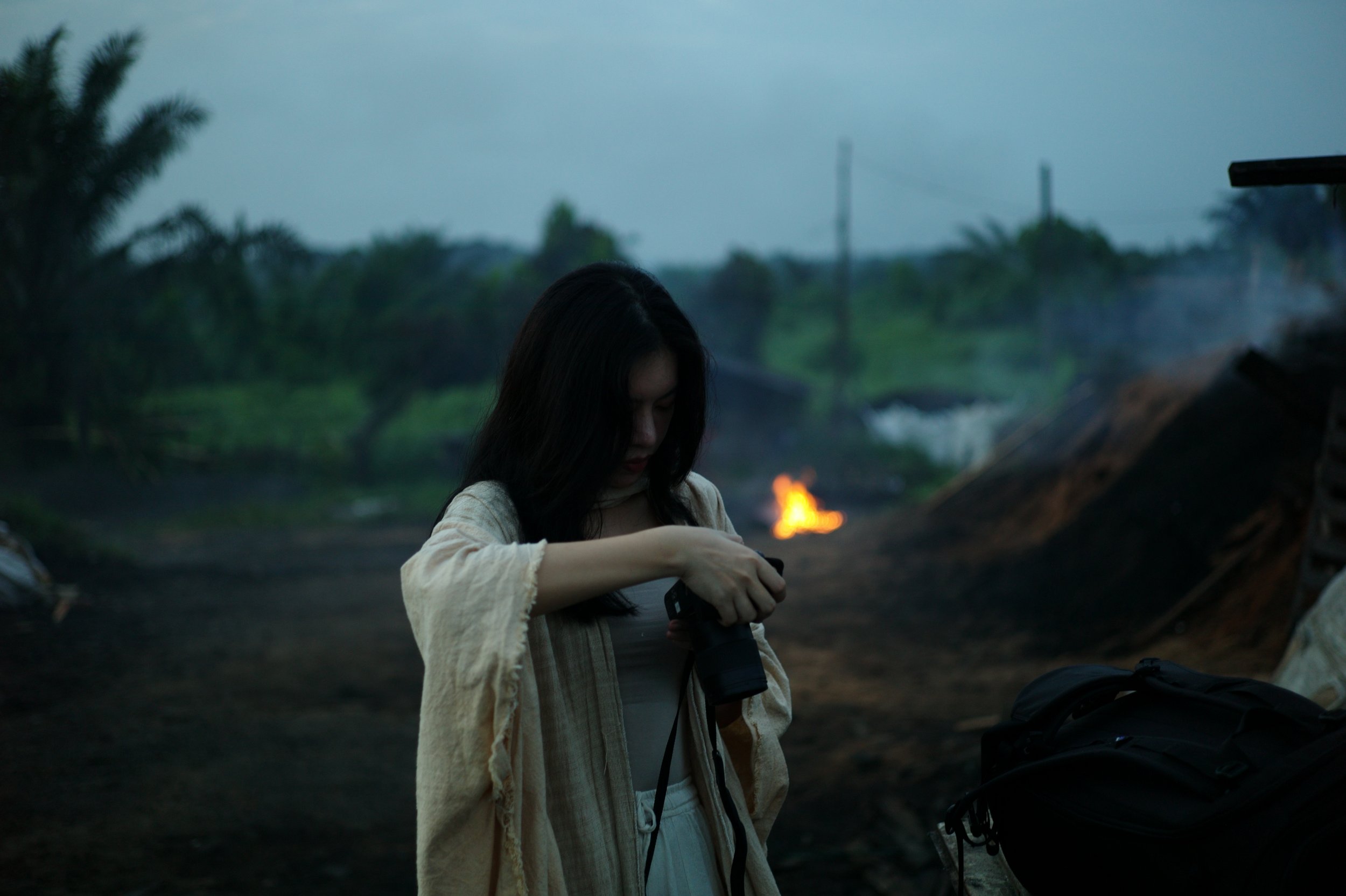A woman with dark hair wearing a beige jacket looks into a camera in a rural outdoor setting with smoke and a small fire burning in the background.
