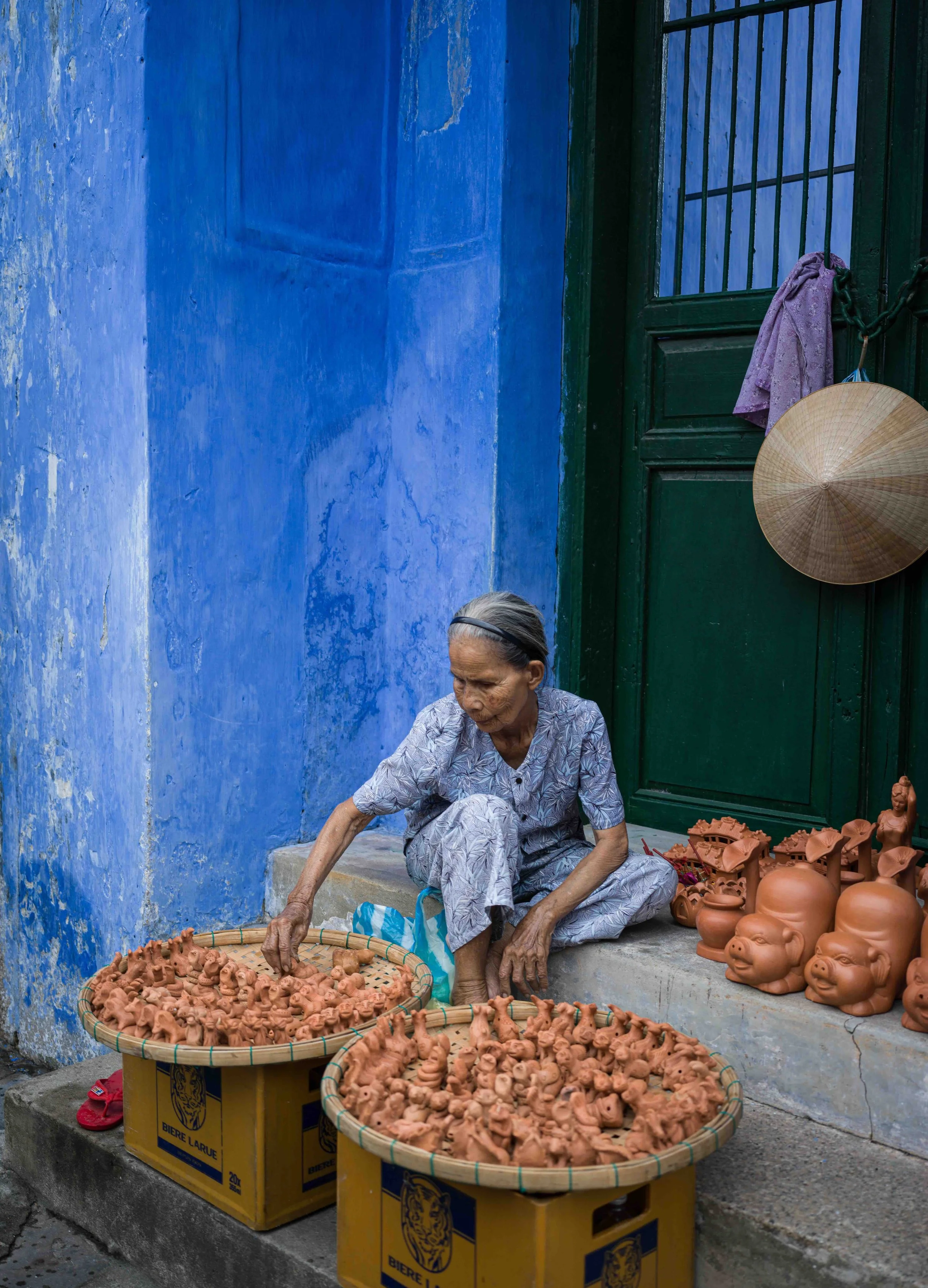 An elderly Vietnamese woman sitting on a stone step outside a house in Hoi An, selling small clay figurines and head sculptures on two large yellow boxes, with a blue wall and a green door in the background.