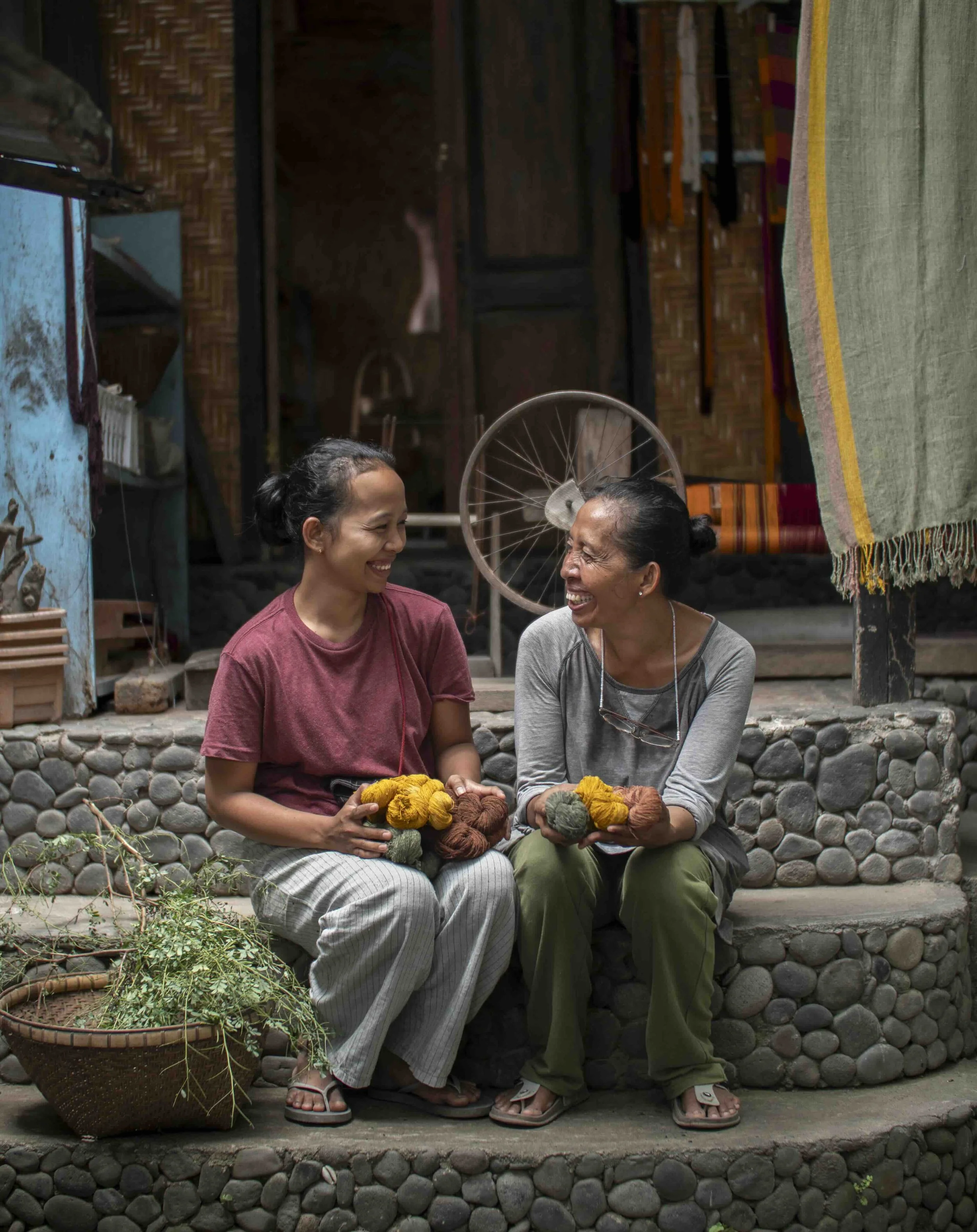 Two Sasak women in Lombok sitting on stone steps, smiling and holding colorful yarn balls, outside a wooden home. They appear to be engaged in a cheerful conversation.