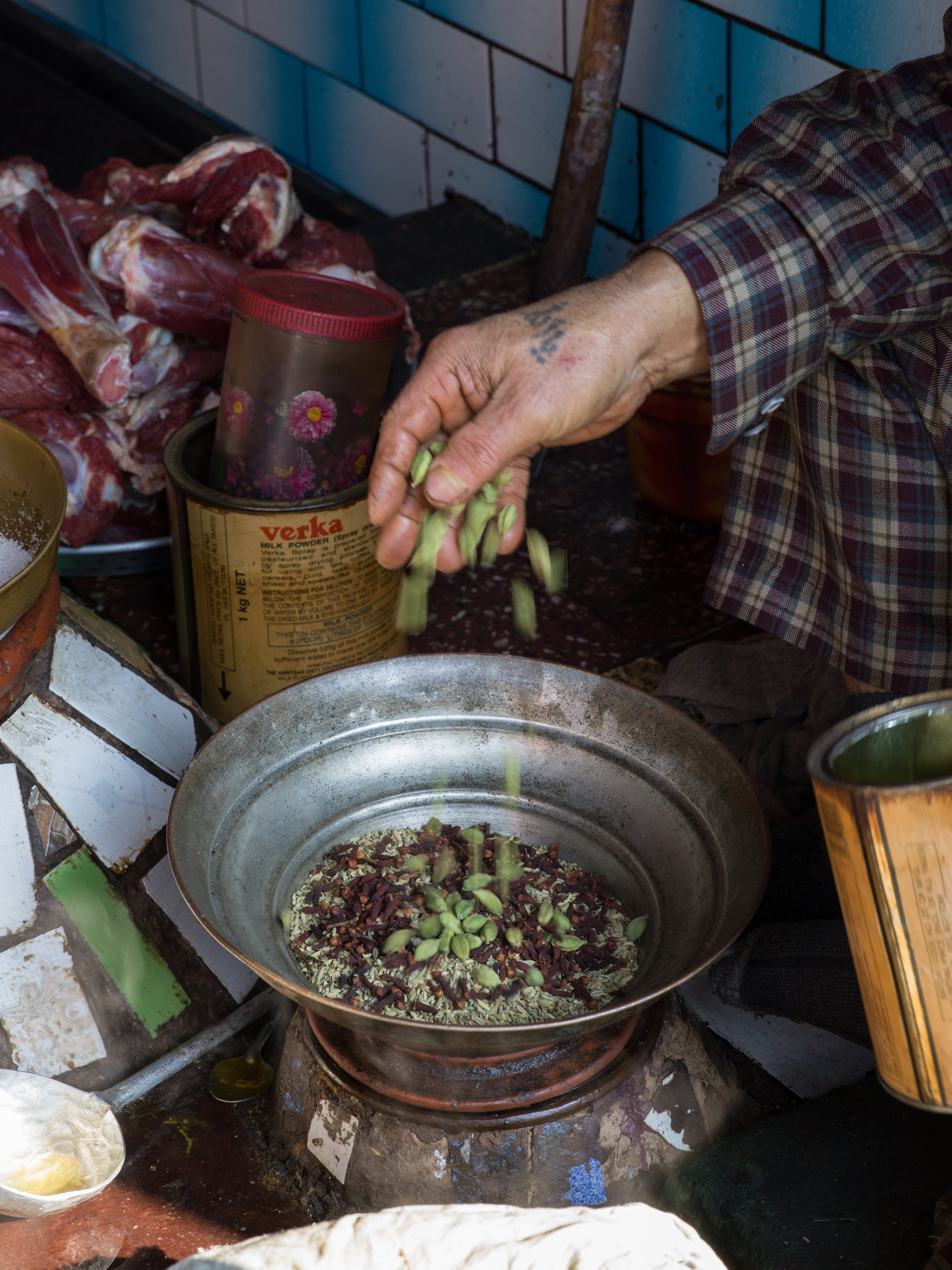 Person adding chopped green herbs into a frying pan on a stove, with raw meat visible in the background.