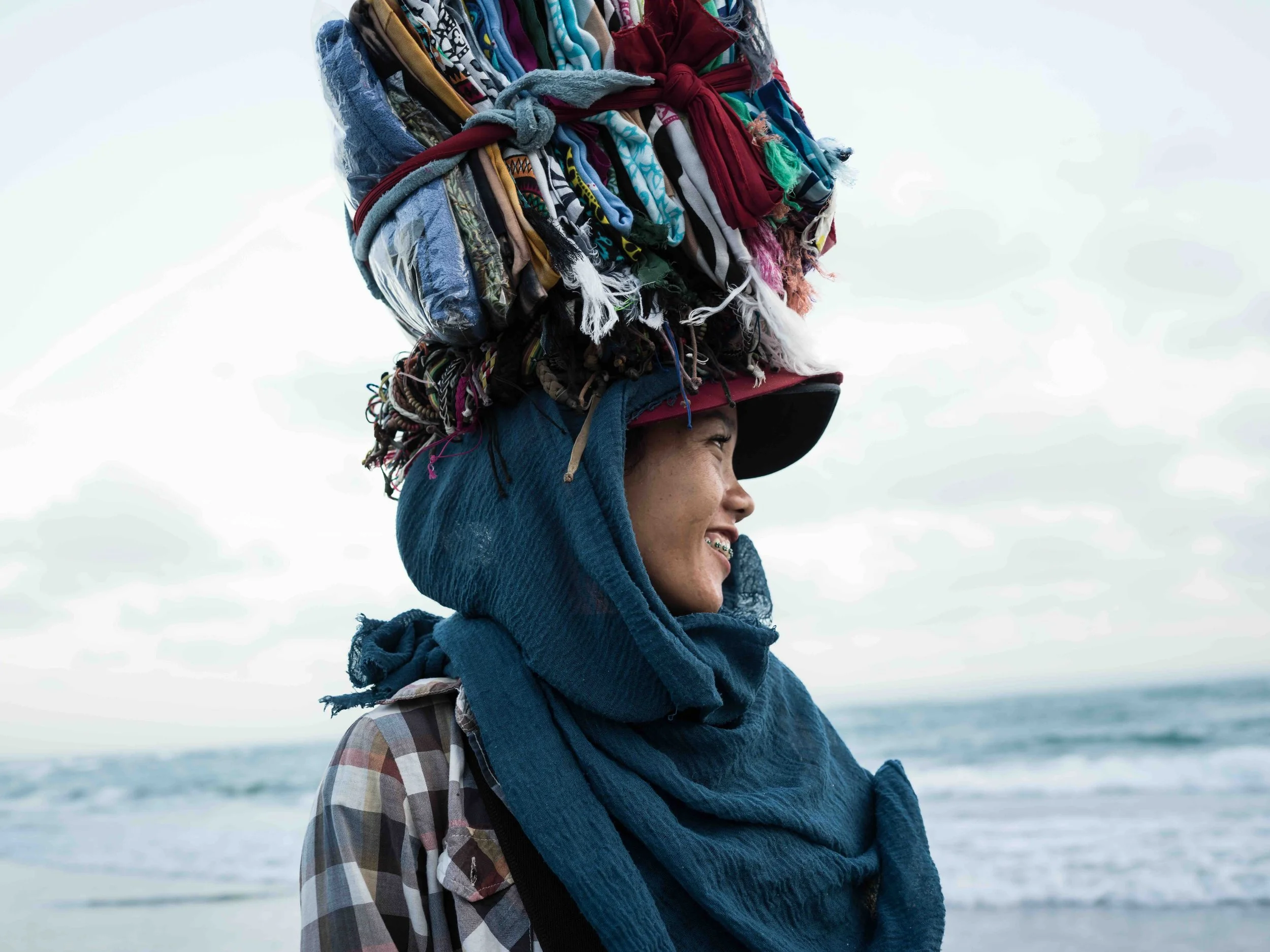 A Balinese young woman at the Canggu beach with a large bundle of colorful fabric tied to her head, smiling and wearing a hoodie and plaid shirt.