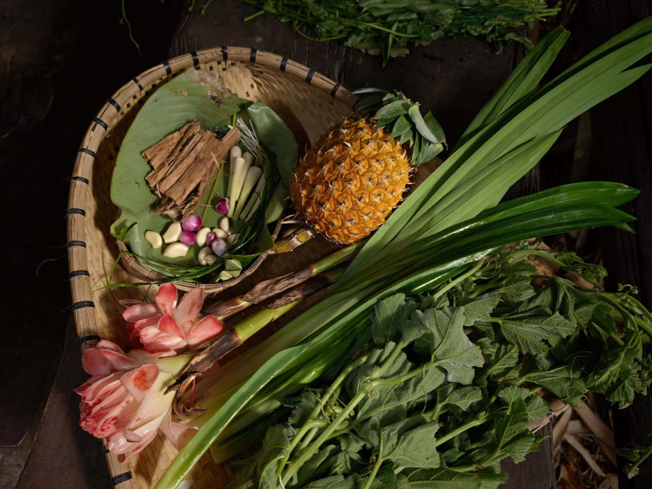 Foraged ingredients for Bidayuh cooking on a woven tapan, including lemongrass, torch ginger flower, garlic, pandan leaves, and pineapple