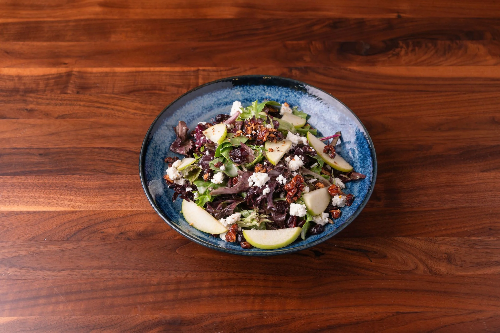 Mixed leafy greens salad with apple slices, candied pecans, blue cheese crumbles, and a dark vinaigrette dressing in a blue bowl on a wooden table.
