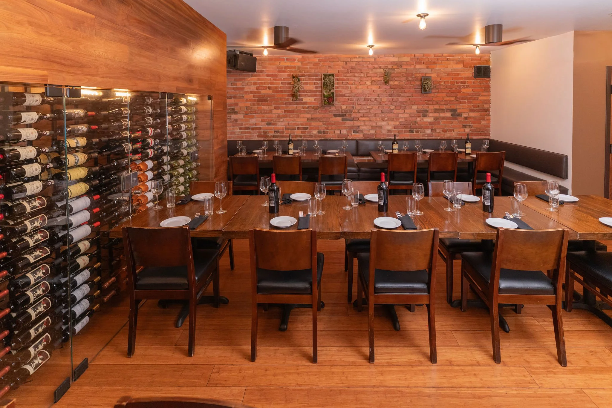 Empty restaurant dining area with wooden tables set with plates, wine glasses, and bottles of wine, surrounded by wooden chairs, brick and wood walls, and wine storage on the left side.