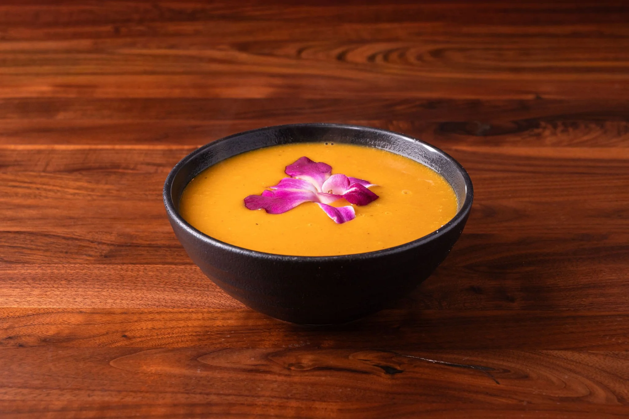 A black bowl filled with yellow-orange soup garnished with pink and purple flower petals on a wooden surface.