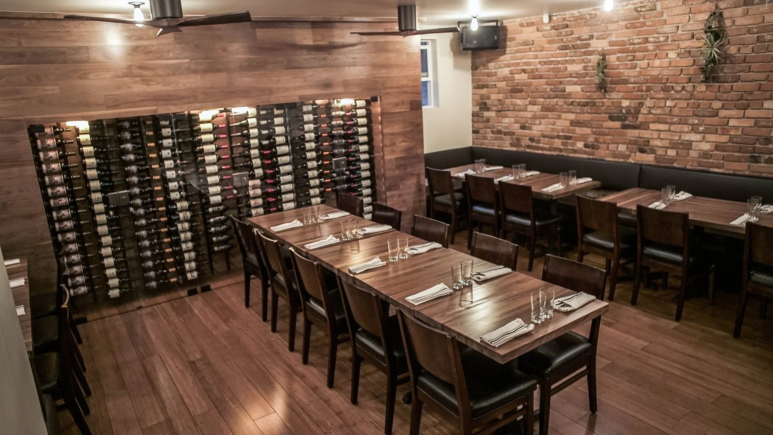 Dining area with a long wooden table set with napkins, glasses, and cutlery, surrounded by wooden chairs, featuring a brick wall with hanging plants and a wine rack on a wooden-paneled wall.