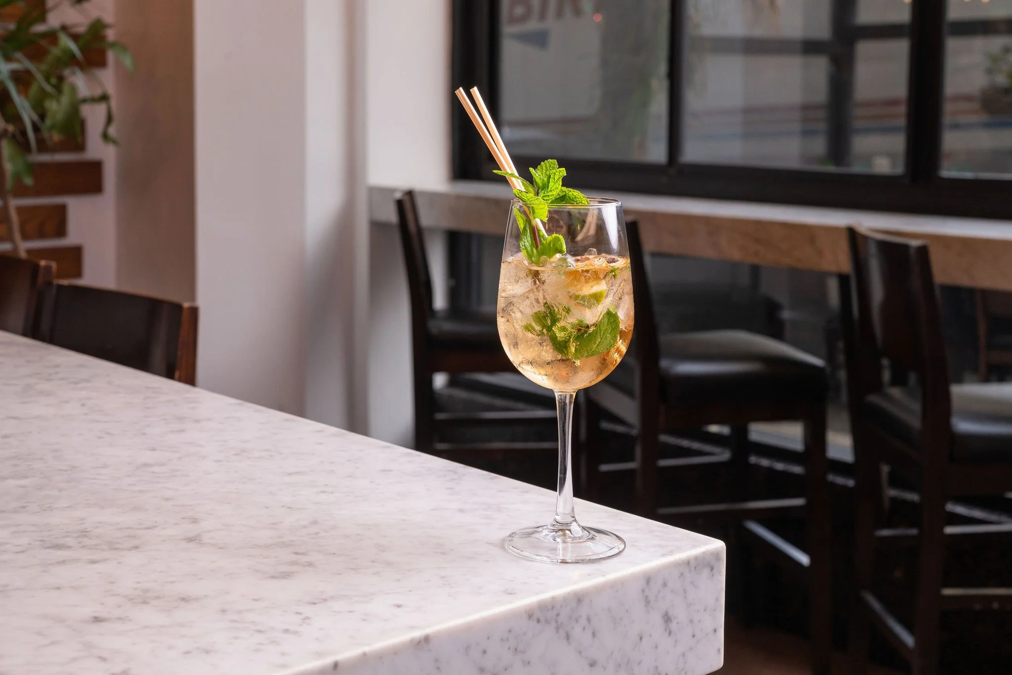 A tall glass of cocktail with mint leaves and straws on a white marble counter in a cafe or restaurant setting.