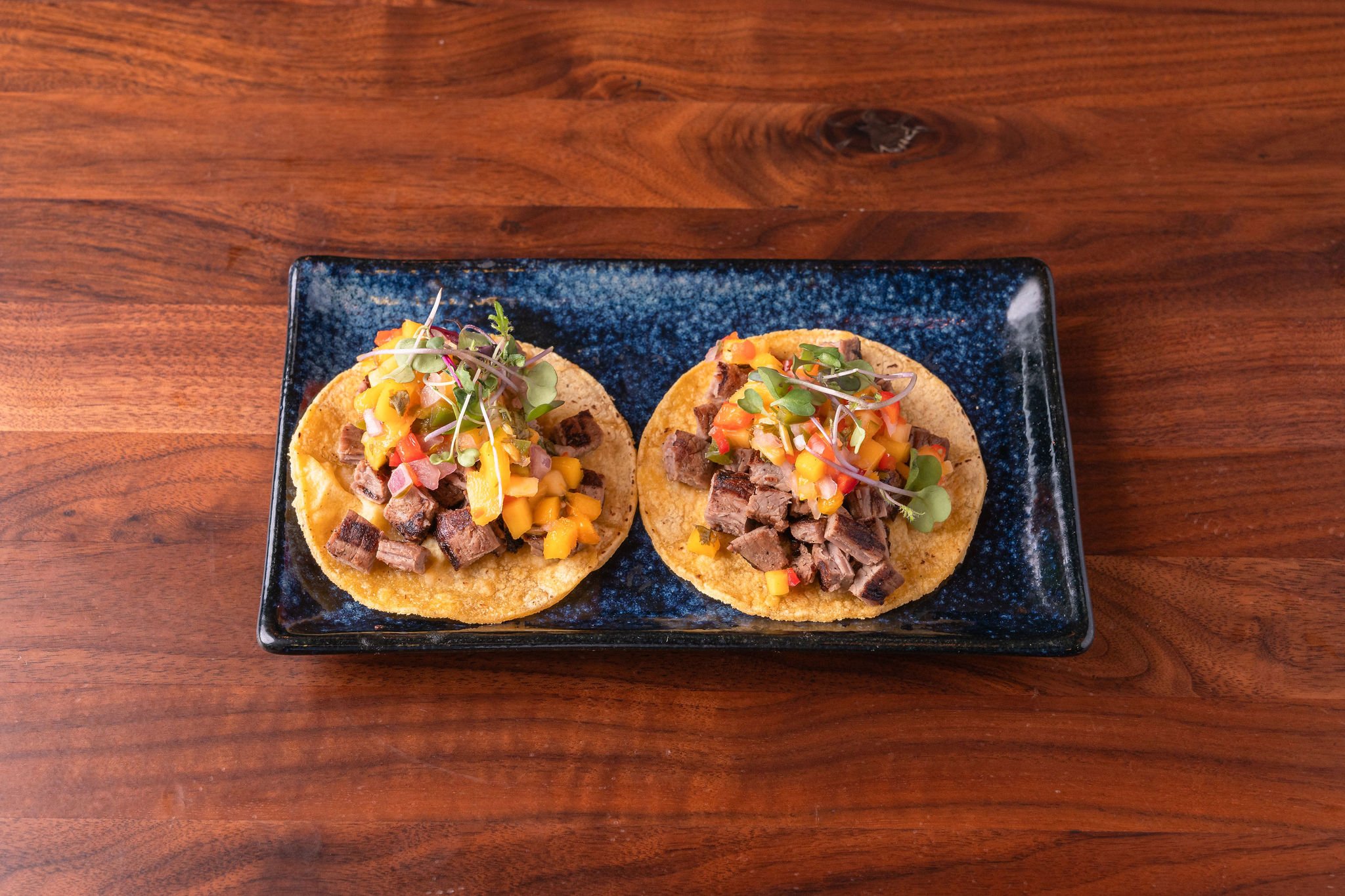 Two tacos topped with diced beef, pineapple, diced red and yellow peppers, and microgreens on a black rectangular plate, set on a wooden table.