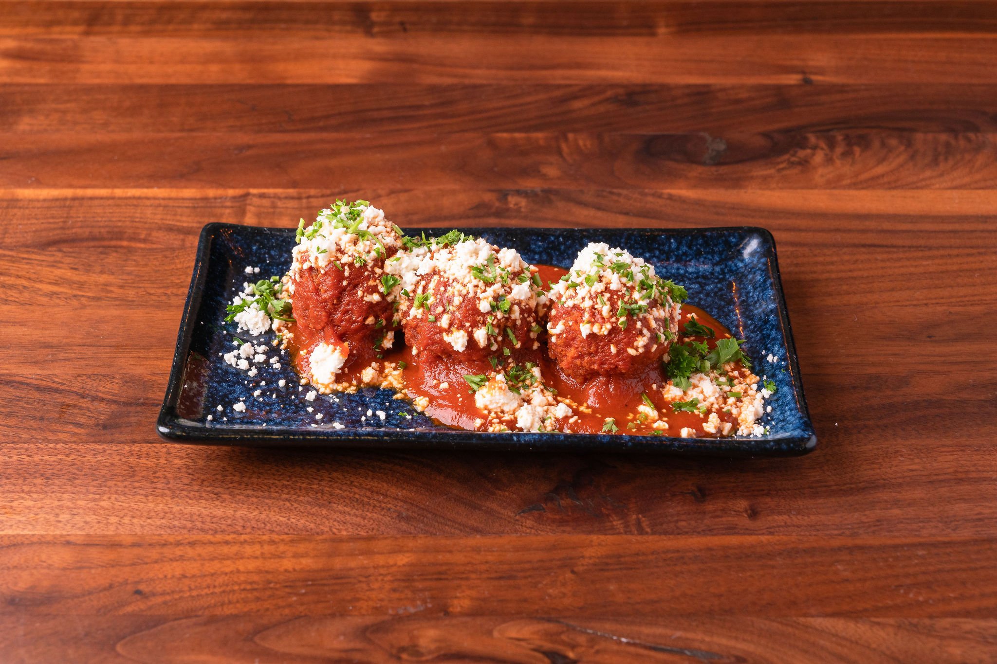 Plate of three meatballs covered in tomato sauce, topped with crumbled cheese and chopped herbs, served on a blue rectangular dish on a wooden table.