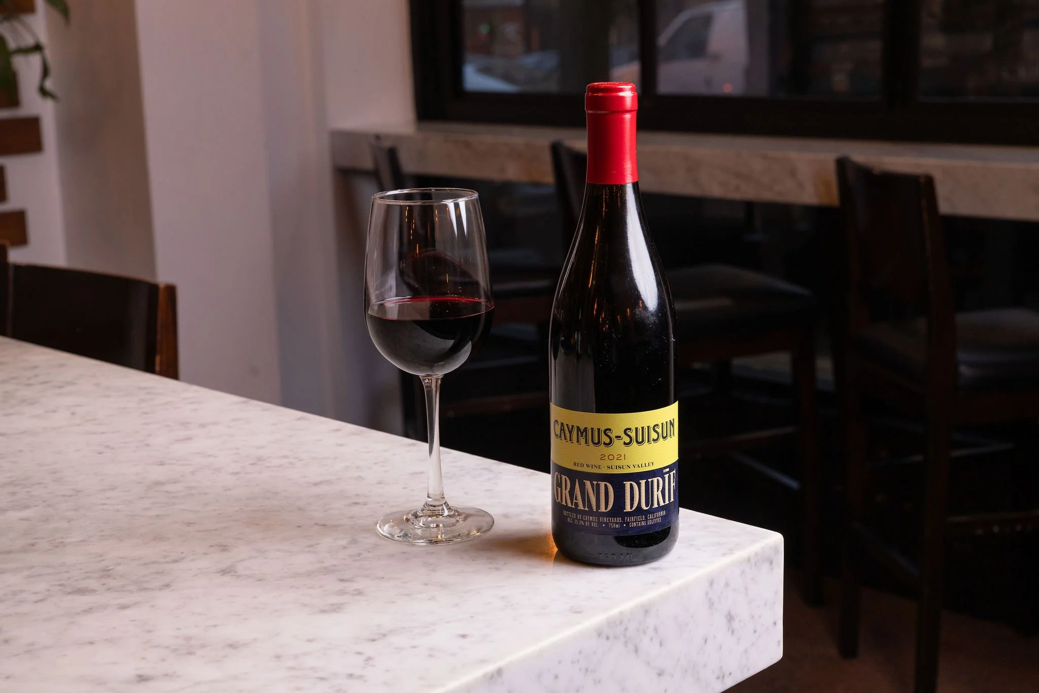 A bottle of red wine labeled 'Caymus-Suisun 2021 Grand Durif' and a glass of red wine on a white marble countertop in a restaurant or cafe setting.
