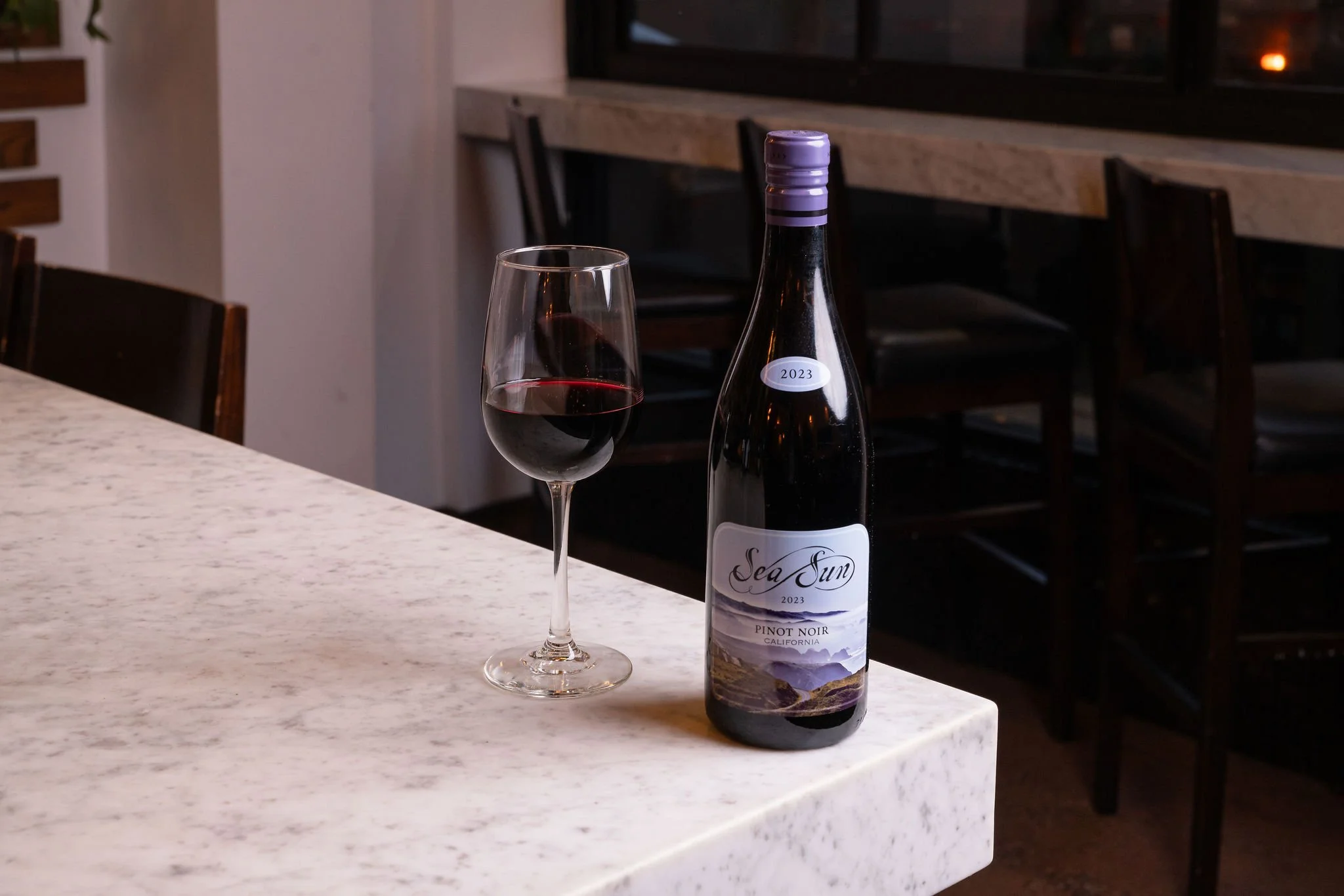 A bottle of Sea Sun 2023 Pinot Noir California wine and a glass of red wine on a light-colored marble countertop in a restaurant or bar setting.