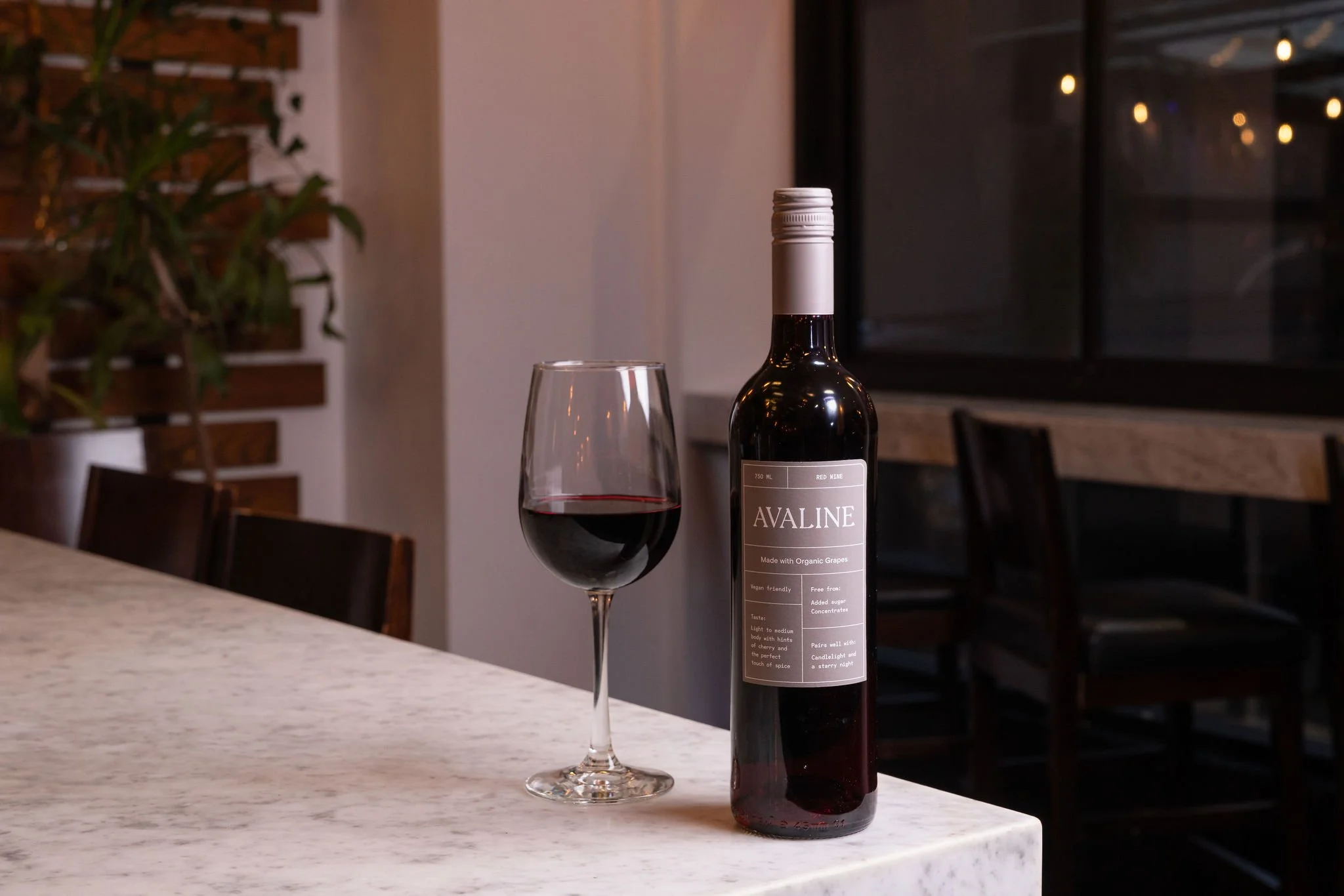 A wine bottle labeled 'Avaline' and a glass of red wine on a white marble countertop in a dimly lit room.