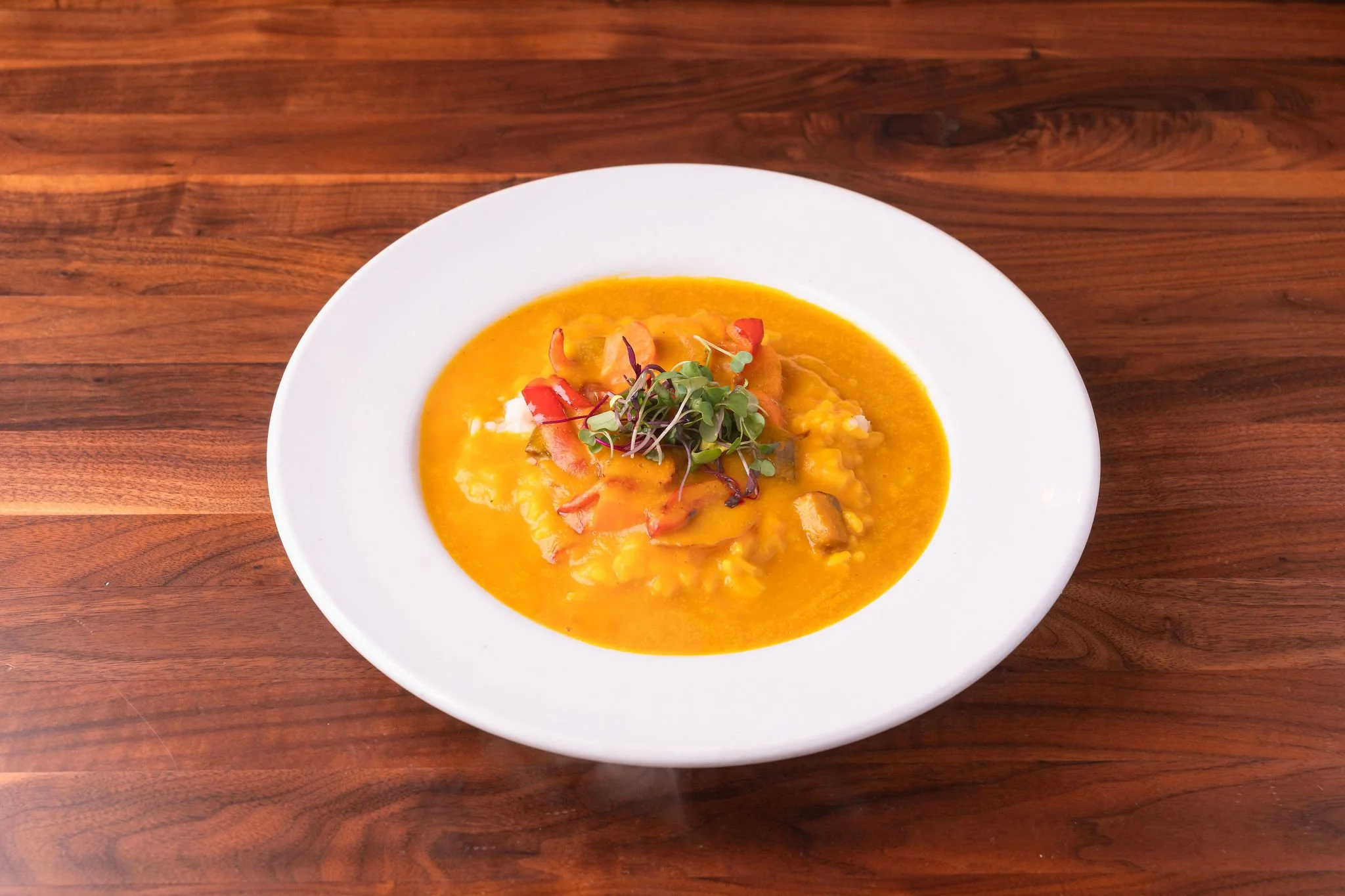 A bowl of pumpkin or squash soup garnished with microgreens and red pepper strips, on a wooden table.