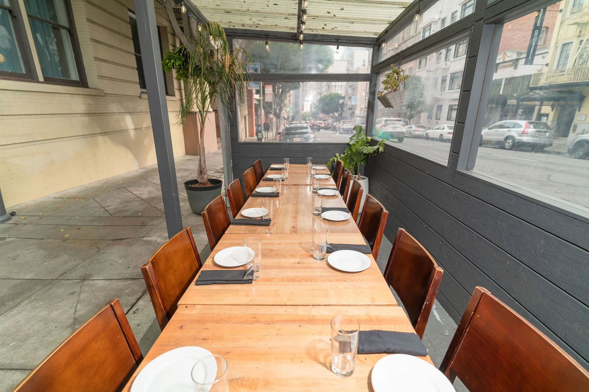 Outdoor dining area with a long wooden table set with white plates, glasses, and black napkins, surrounded by wooden chairs, enclosed with glass panels, with plants and city street view outside.