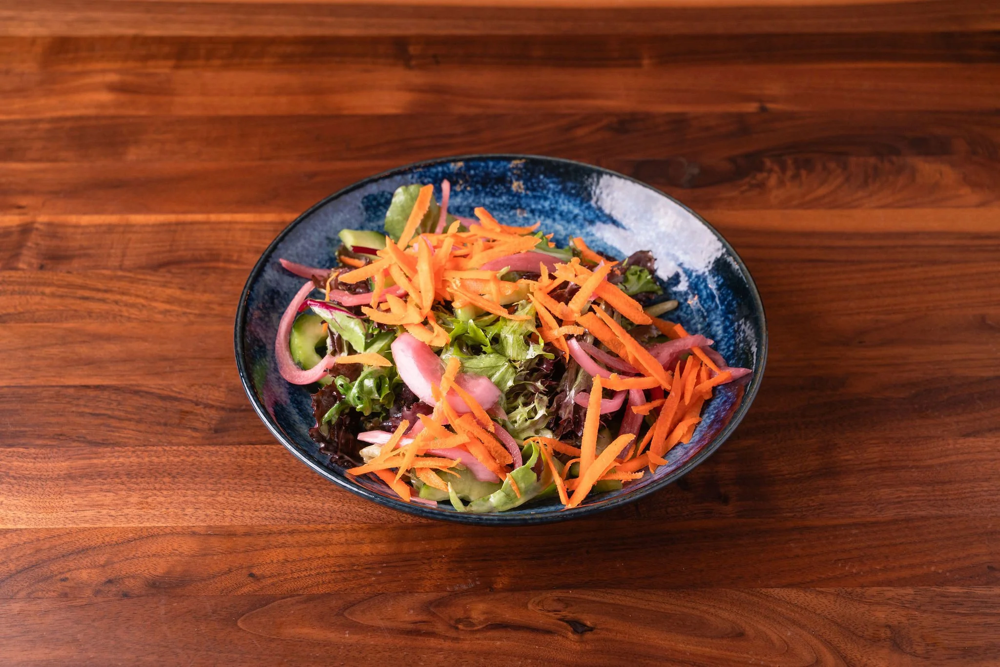 A bowl of mixed salad with shredded carrots, sliced cucumbers, red onions, and leafy greens on a wooden table.