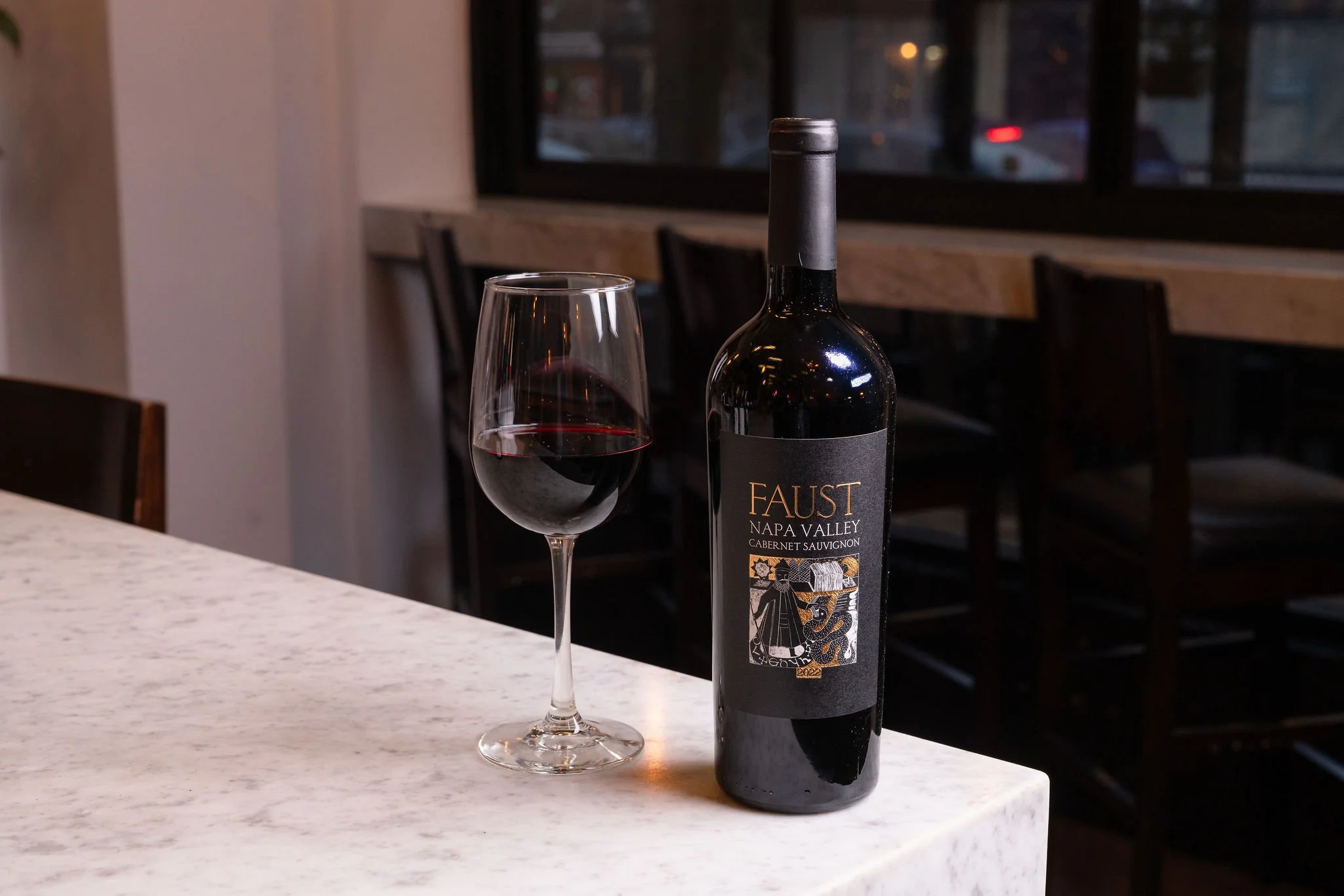 A bottle of Faust Napa Valley Cabernet Sauvignon and a glass of red wine on a white marble table in a dimly lit restaurant with wooden chairs and a window in the background.