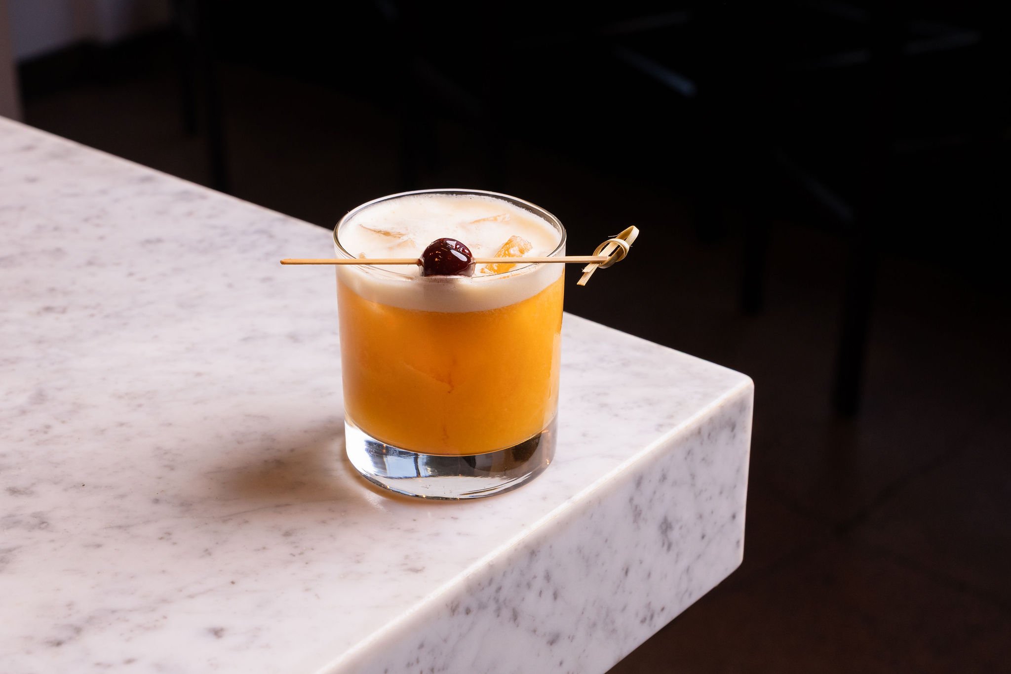 Cocktail with orange-colored liquid topped with foam and garnished with a cherry on a skewer on a white marble surface.