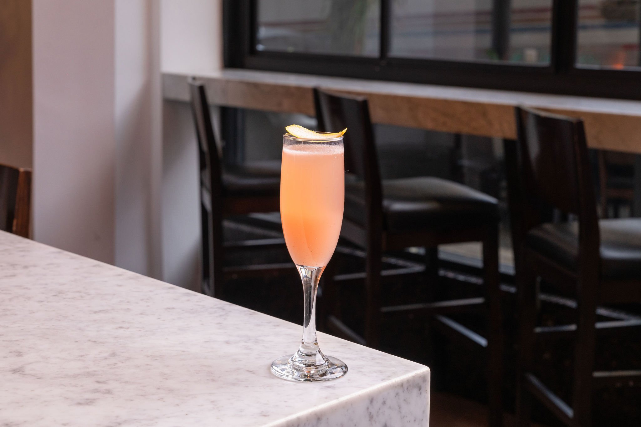 A pink cocktail in a tall champagne flute garnished with a lemon slice, placed on a white marble-countertop inside a restaurant or cafe.