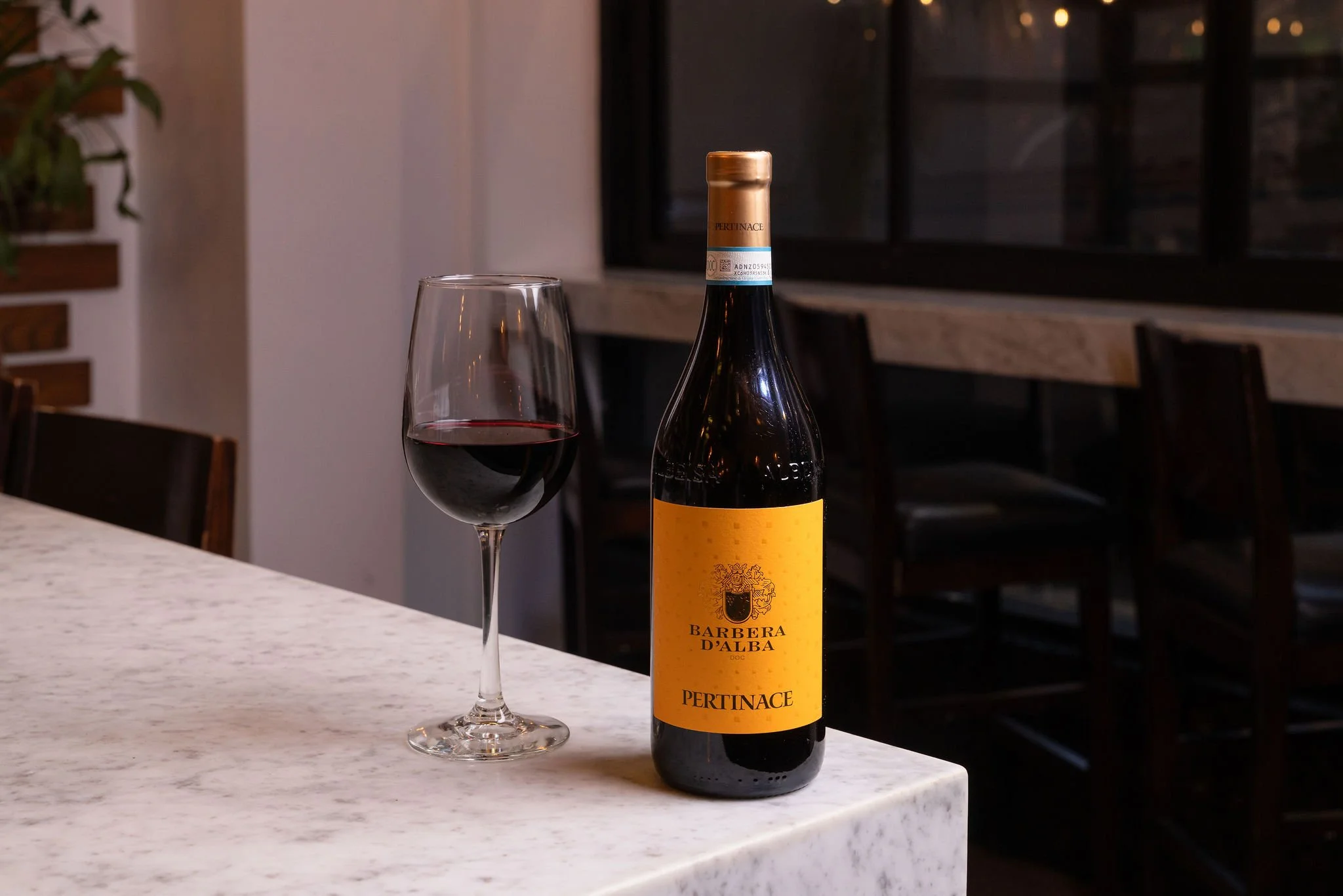 A bottle of red wine labeled 'Barbera D'Alba' with a glass of red wine beside it, on a white marble countertop in a dimly lit room with dark chairs and a window in the background.