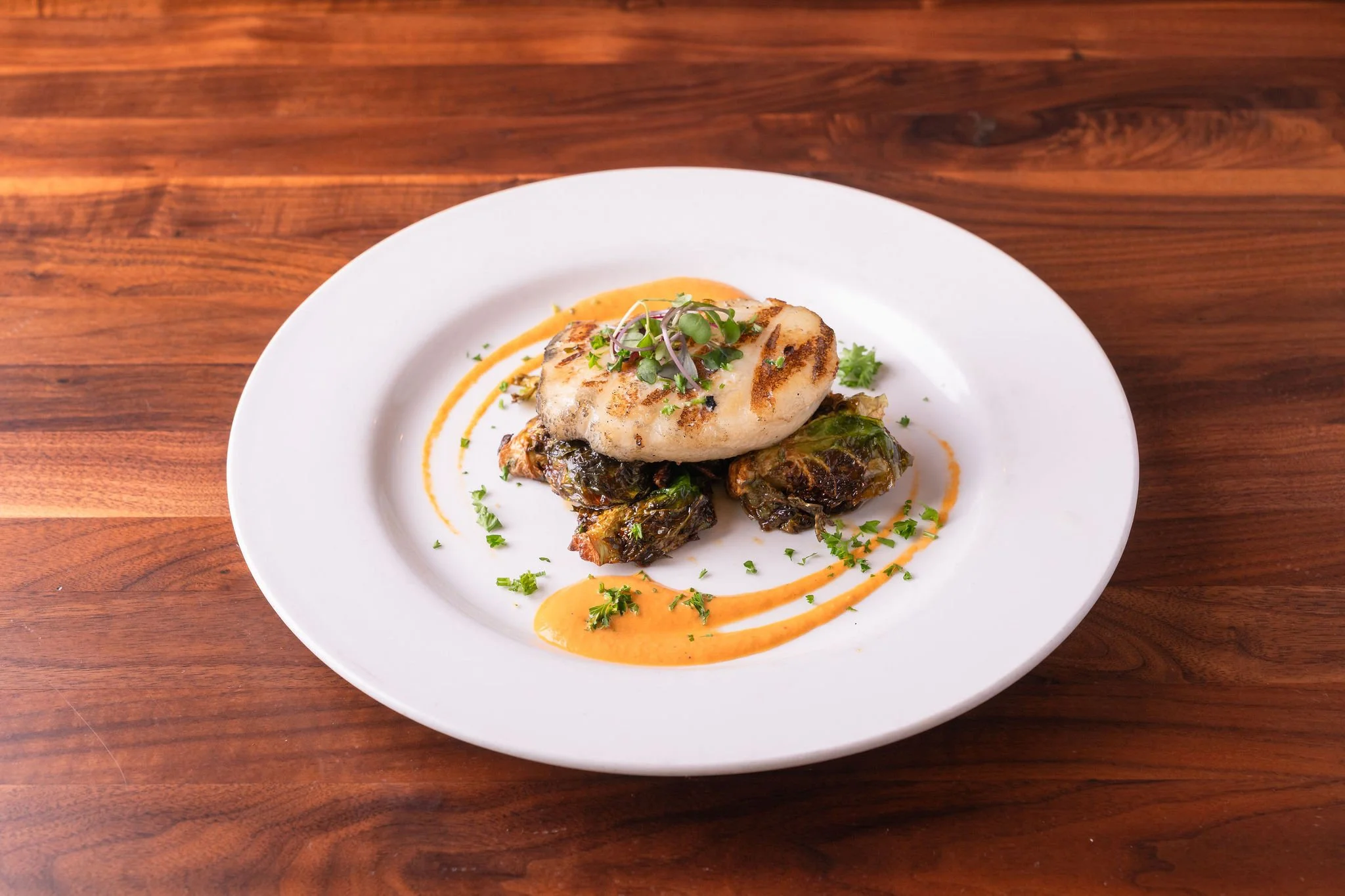 Grilled fish fillet on a bed of roasted vegetables with orange sauce and microgreens on a white plate.