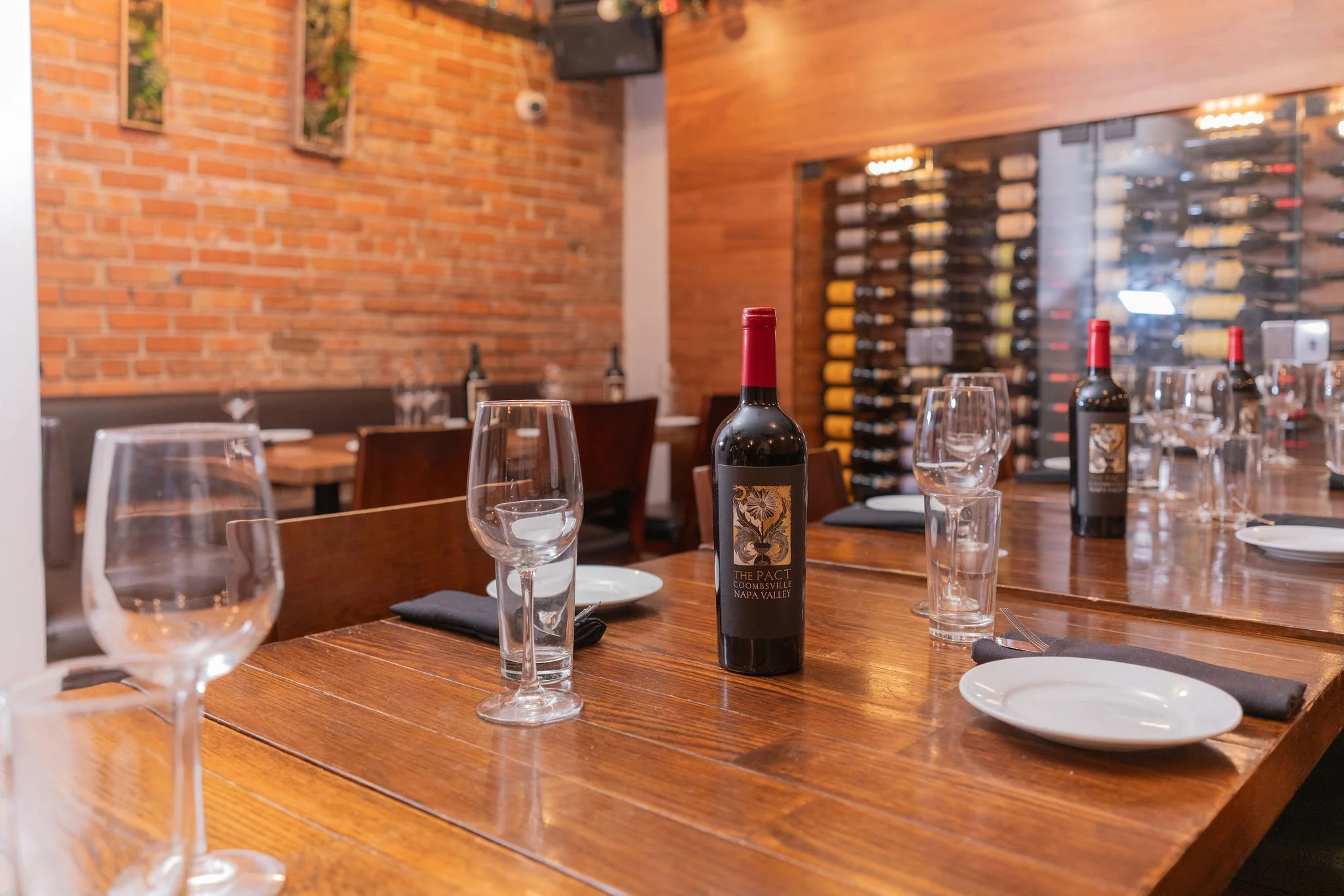 A restaurant table set for wine tasting with glasses, plates, napkins, and bottles of wine. The background features a brick wall and a wine rack filled with bottles.