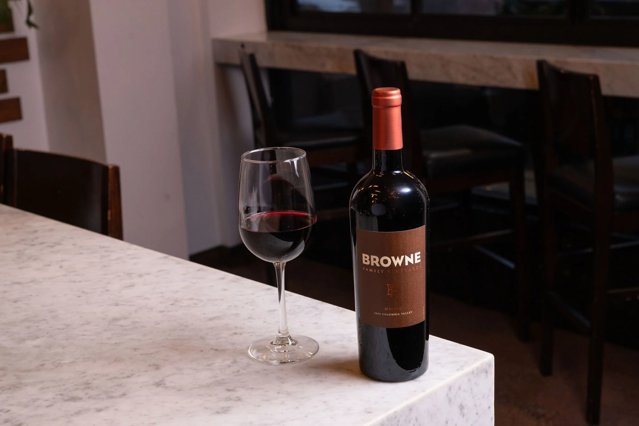 A bottle of red wine labeled "Browne" and a glass of red wine on a white marble counter with dark wooden chairs in the background.