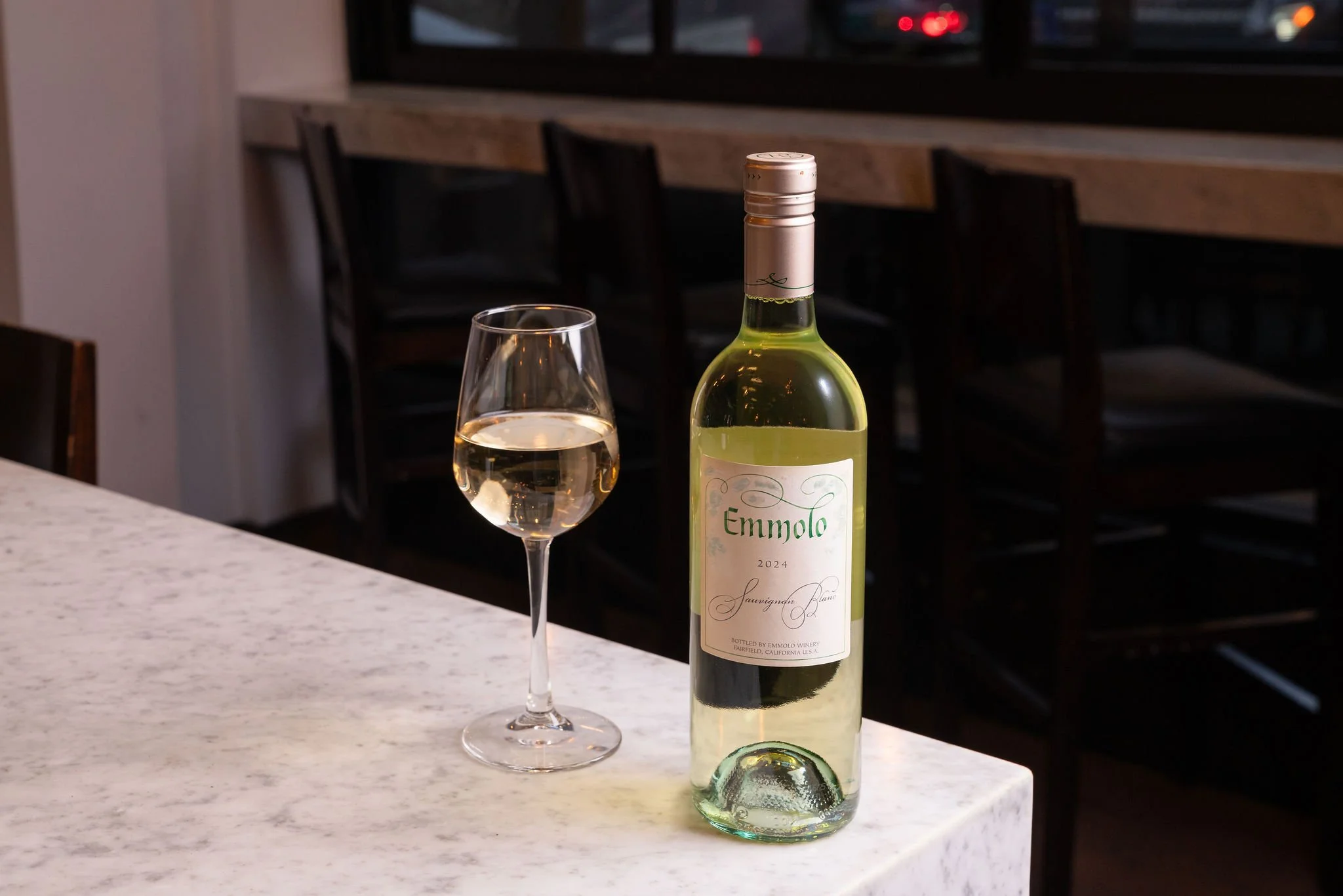 A bottle of Emmolo Sauvignon Blanc wine and a glass of white wine on a white marble countertop in a dimly lit bar or restaurant setting.