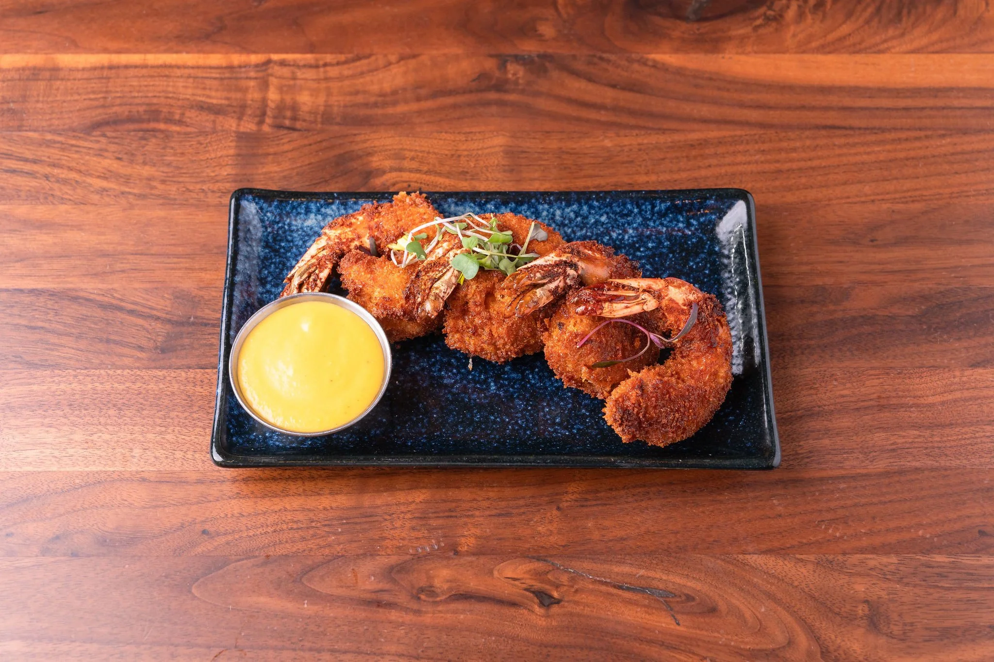 Fried shrimp with microgreens garnishing, served with a yellow dipping sauce on a blue plate on a wooden table.
