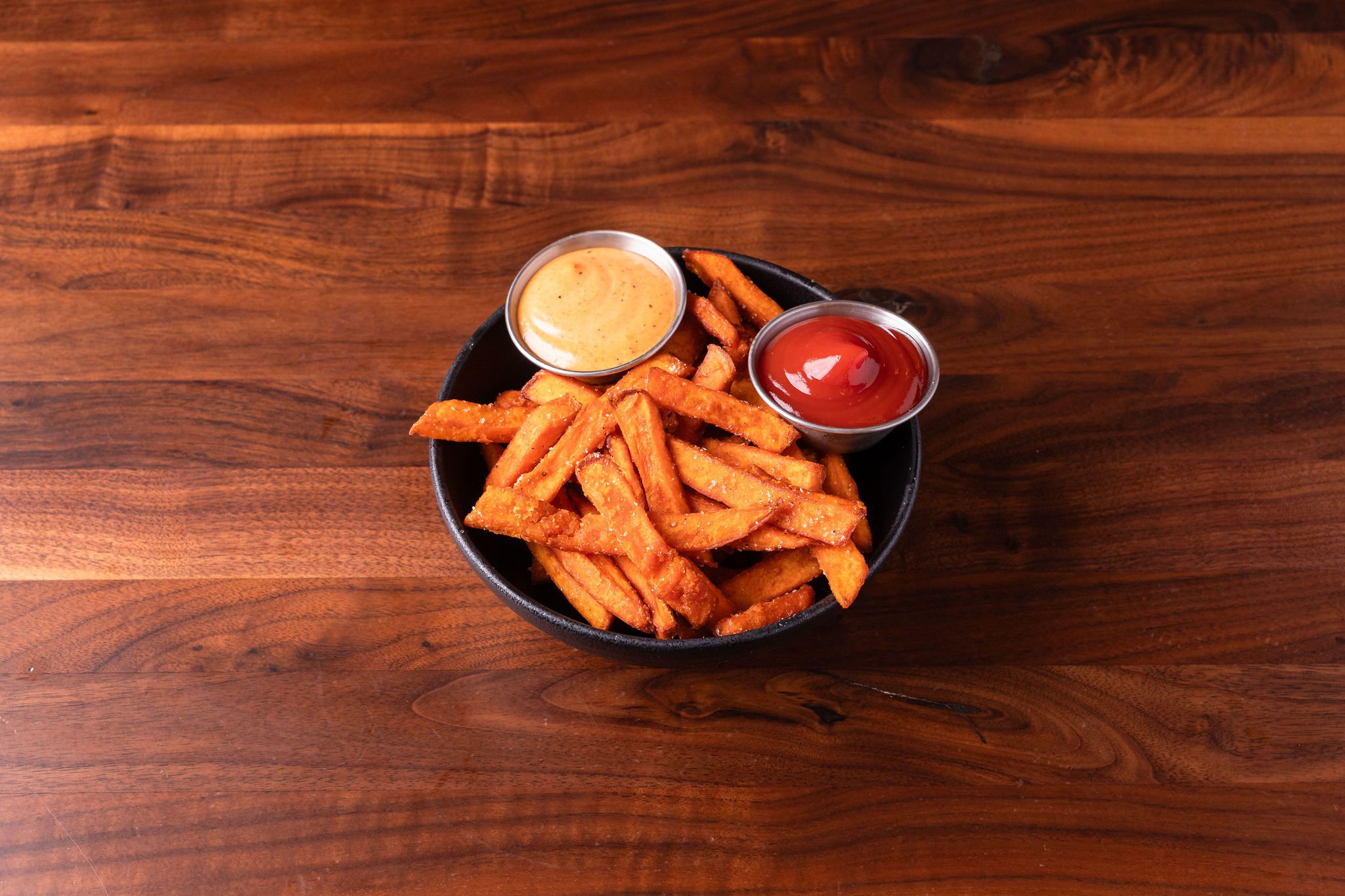 Basket of crispy French fries with two small cups of dipping sauces, one with ketchup and the other with a spicy mayo, on a wooden table.