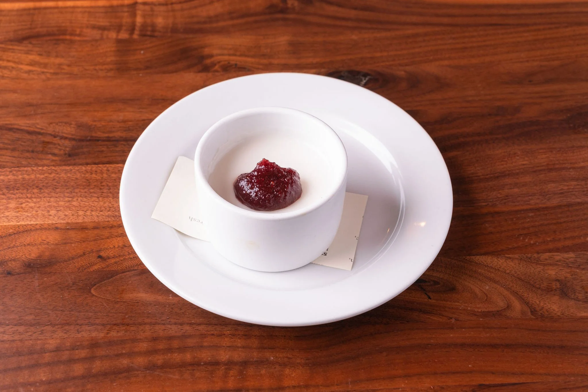 White cup with red jam or jelly on top, placed on a white plate on a wooden table.