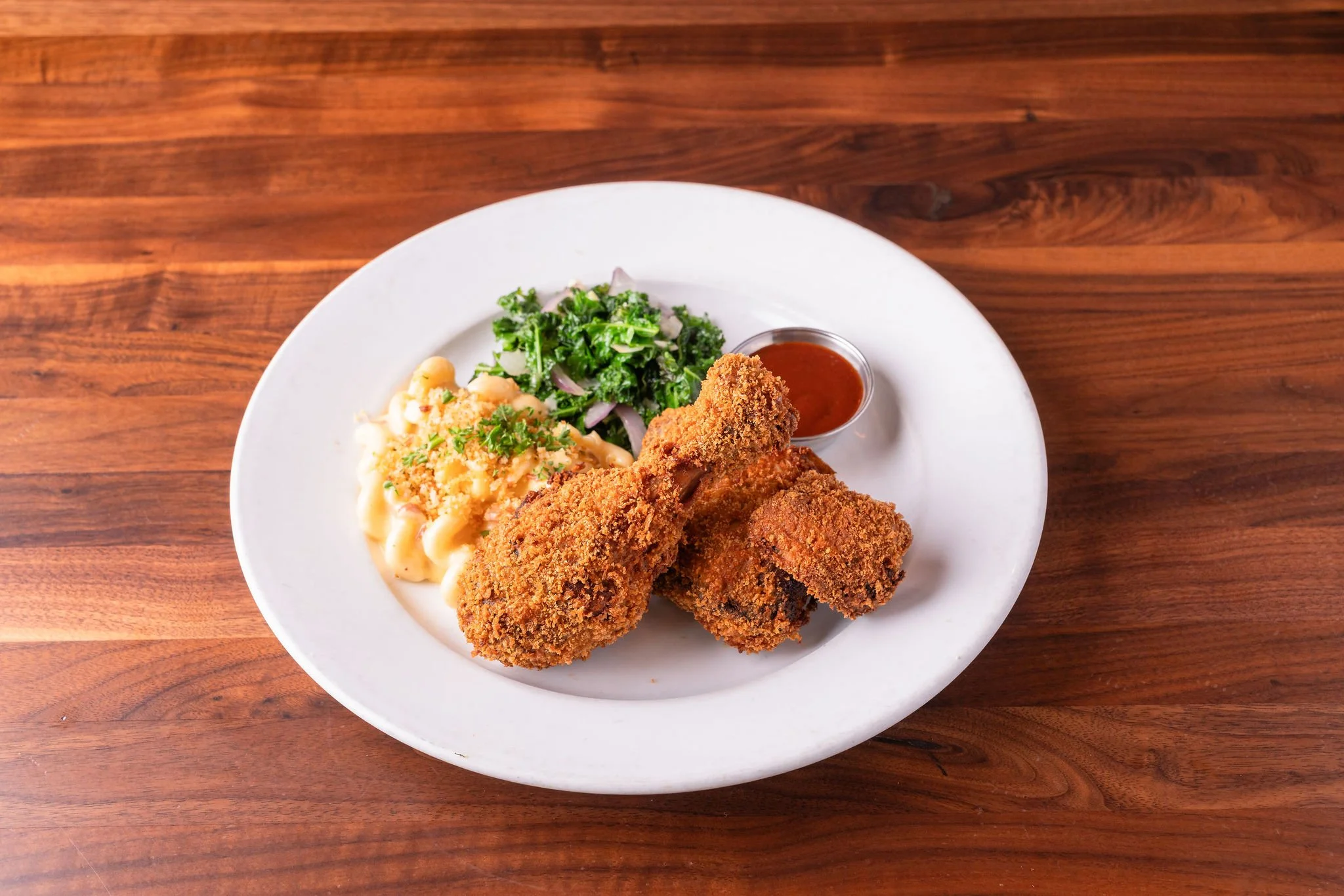 Fried chicken drumstick and square pieces with side salad, macaroni and cheese, and a small cup of red sauce on a white plate on a wooden table.