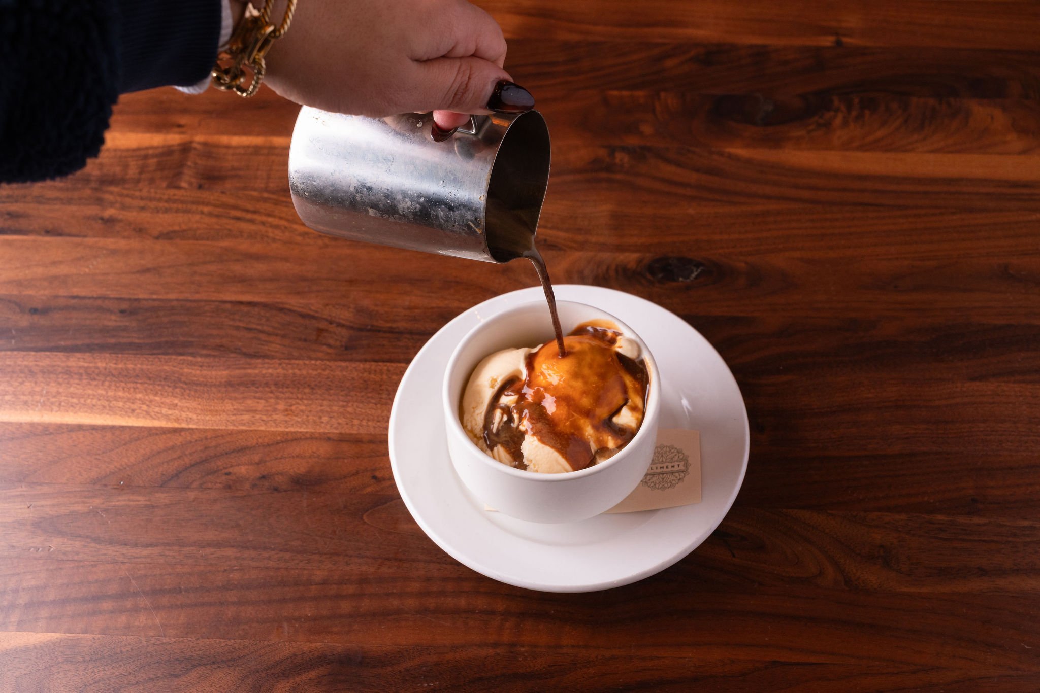 A hand holding a metal pitcher pouring coffee over a bowl of ice cream on a wooden table.