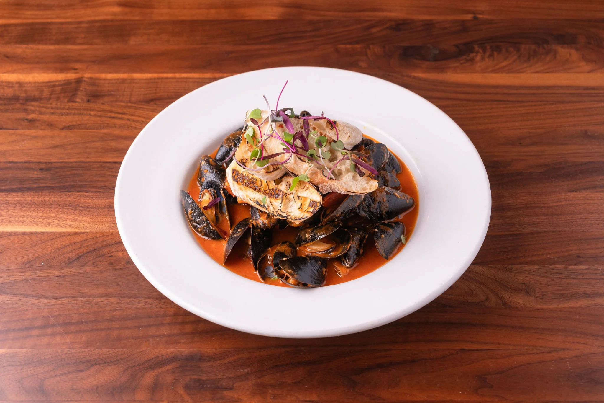 A white bowl of seafood pasta with boat-shaped shells and grilled chicken, garnished with microgreens on top, all served in a tomato sauce on a wooden table.