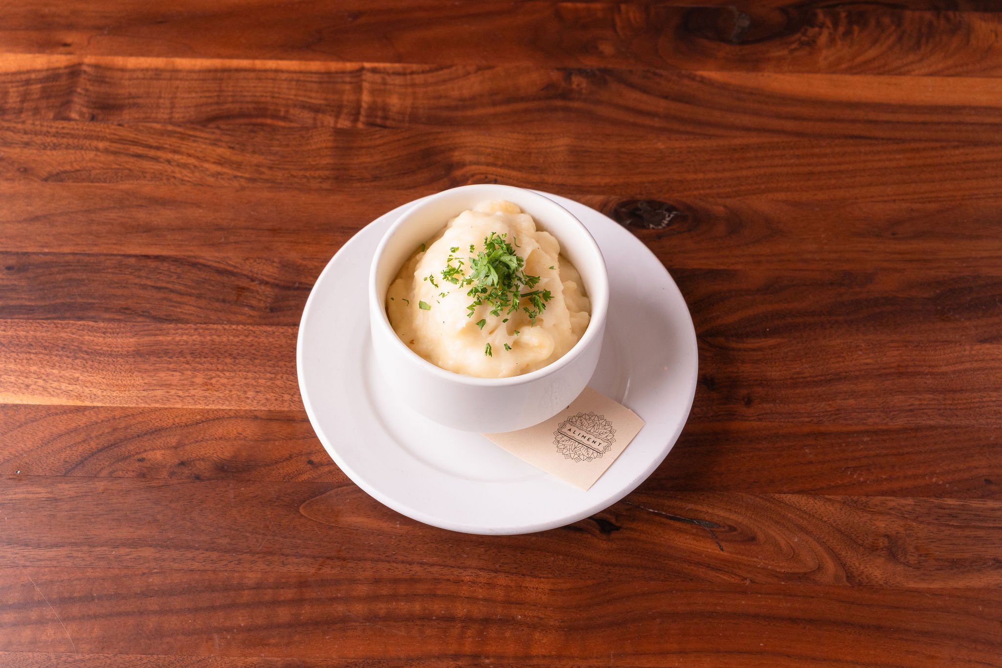 Creamy mashed potatoes topped with chopped parsley served in a white bowl on a white plate with a small paper label underneath, on a wooden table.