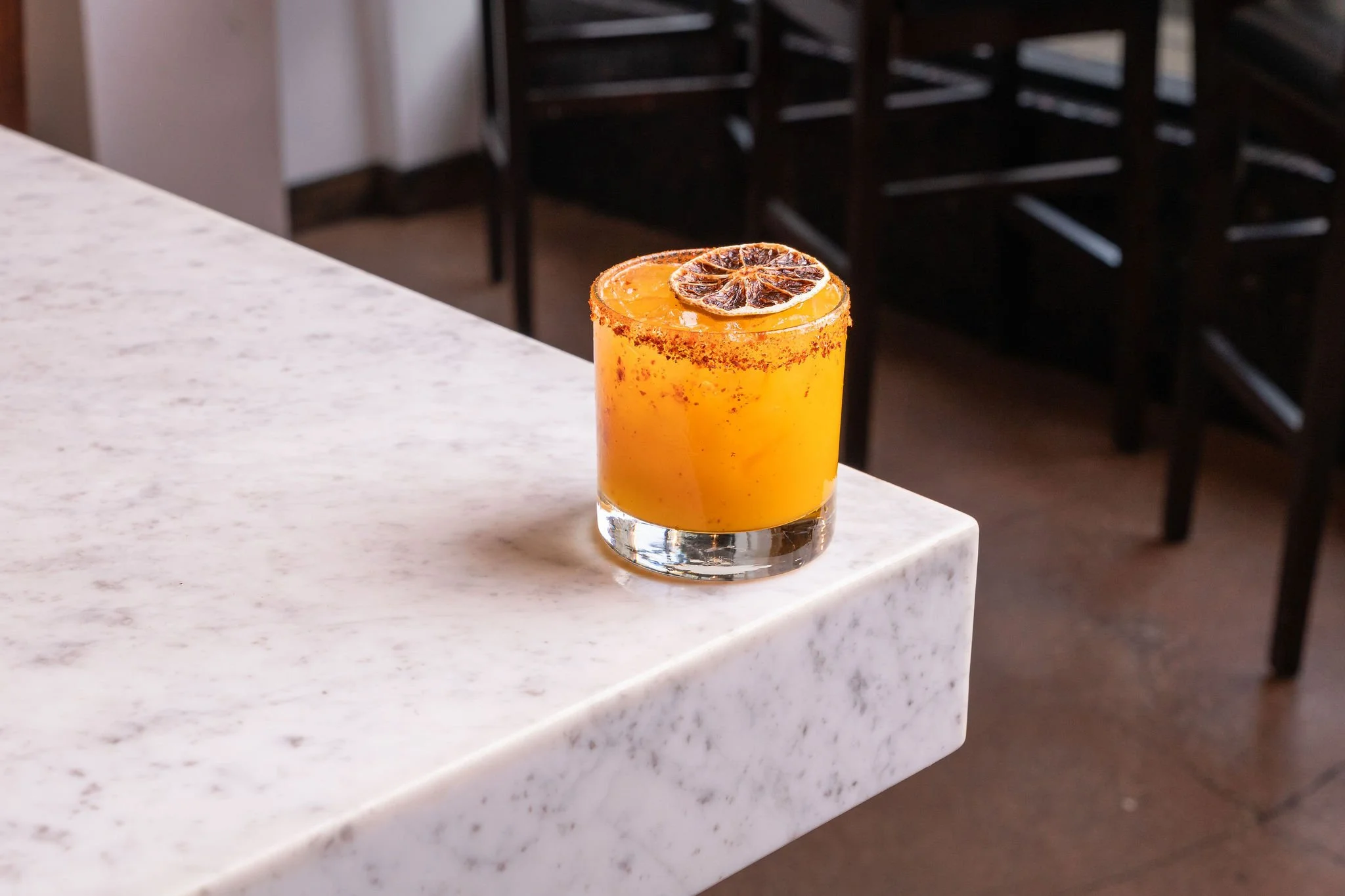 A glass of orange cocktail with a dried lime wheel garnish, placed on a white marble countertop in a restaurant or bar.