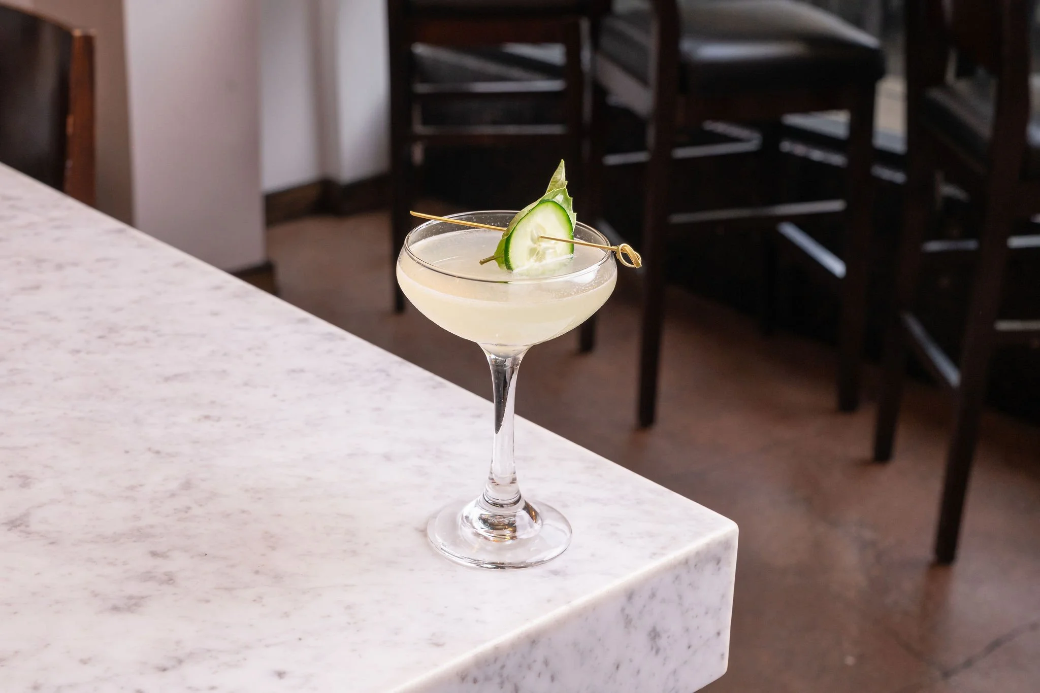 A cocktail glass with a pale yellow drink, garnished with a cucumber slice and a cucumber leaf, placed on a white marble counter in a restaurant or bar setting.