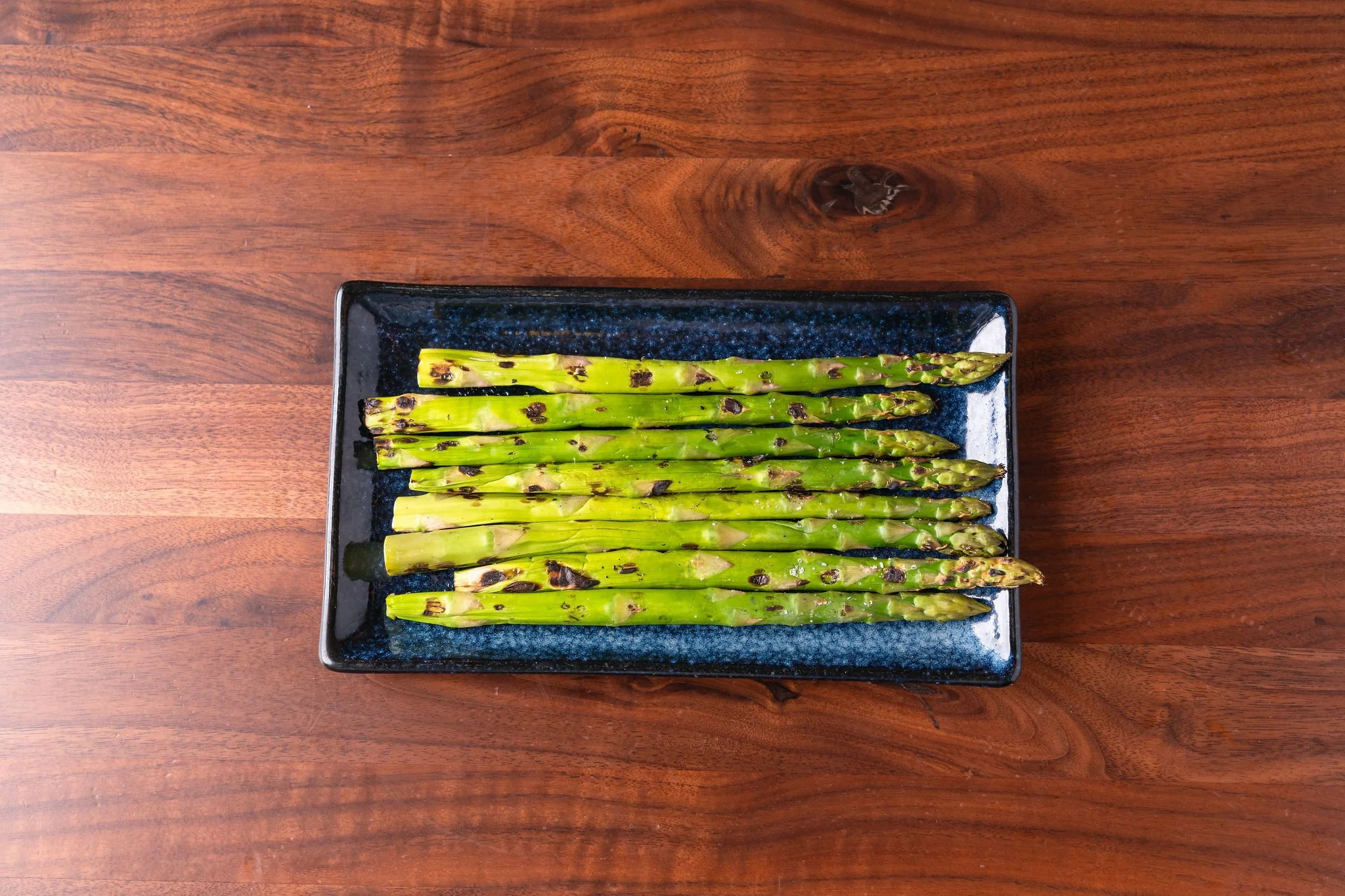 A rectangular black ceramic plate with charred grilled asparagus spears on a wooden table.