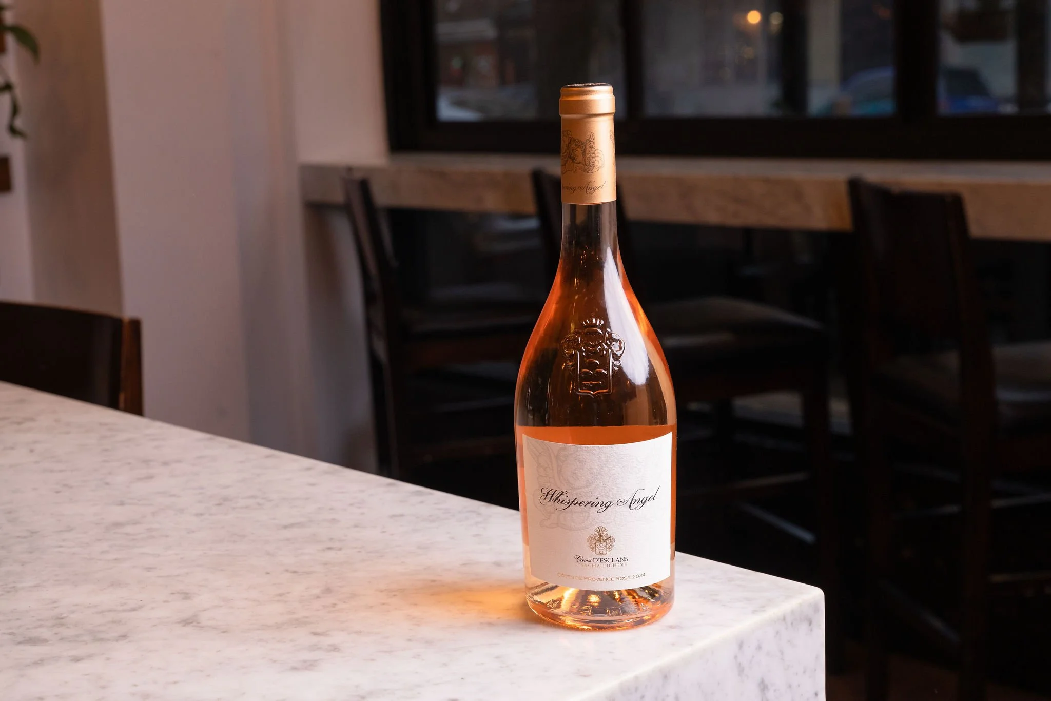 A bottle of Whispering Angel rosé wine on a white marble counter in a restaurant with dark chairs and large windows in the background.
