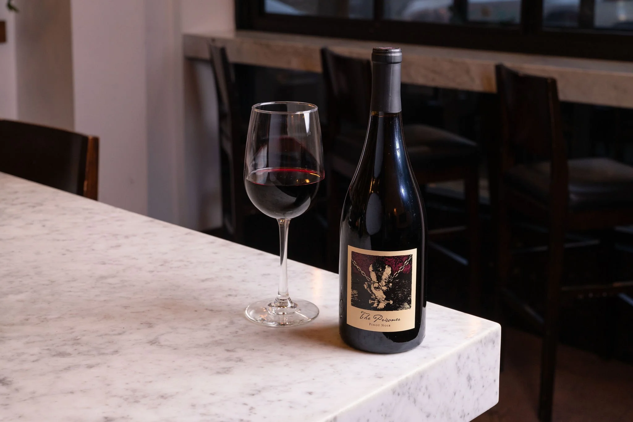 A bottle of Pinot Noir wine labeled 'The Prisoner' and a glass of red wine on a white marble countertop in a restaurant setting.