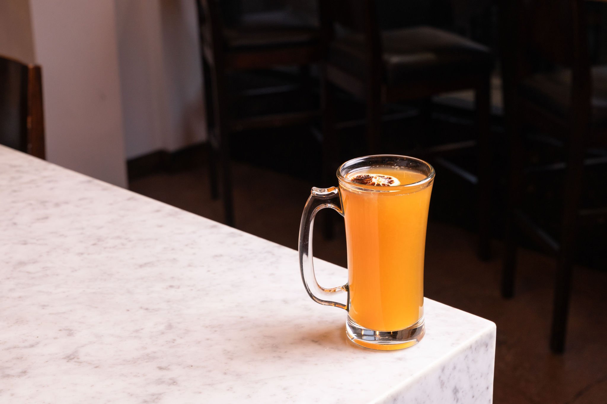 A tall glass of orange-colored drink with a slice of dried orange on top, placed on a white marble countertop in a cafe or restaurant setting.