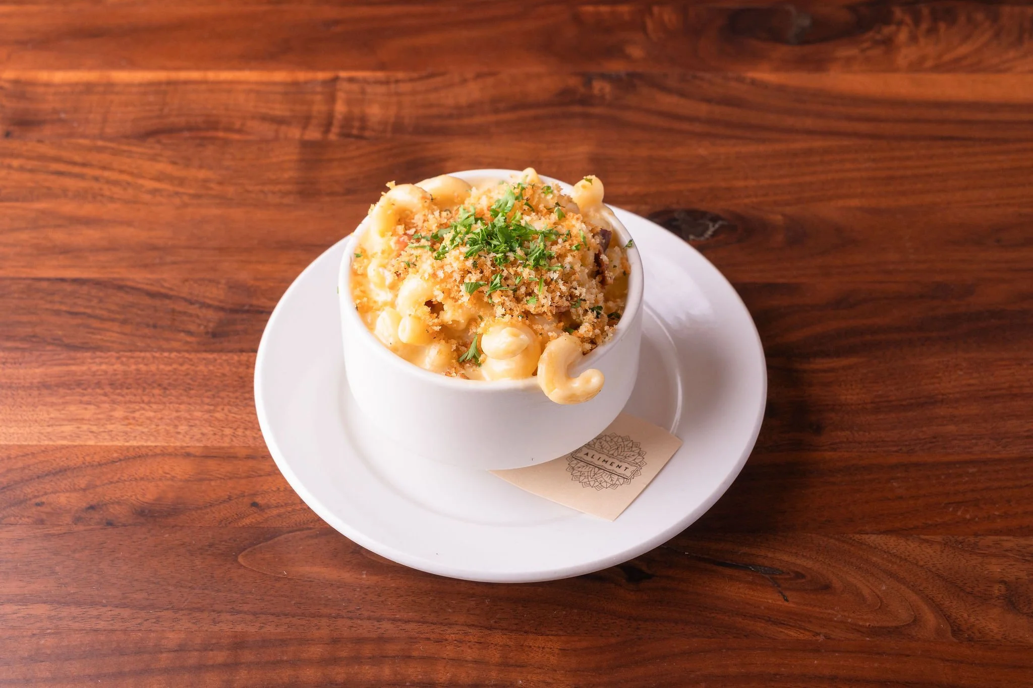 Creamy macaroni and cheese topped with bread crumbs and parsley, served in a white bowl on a white plate on a wooden table.