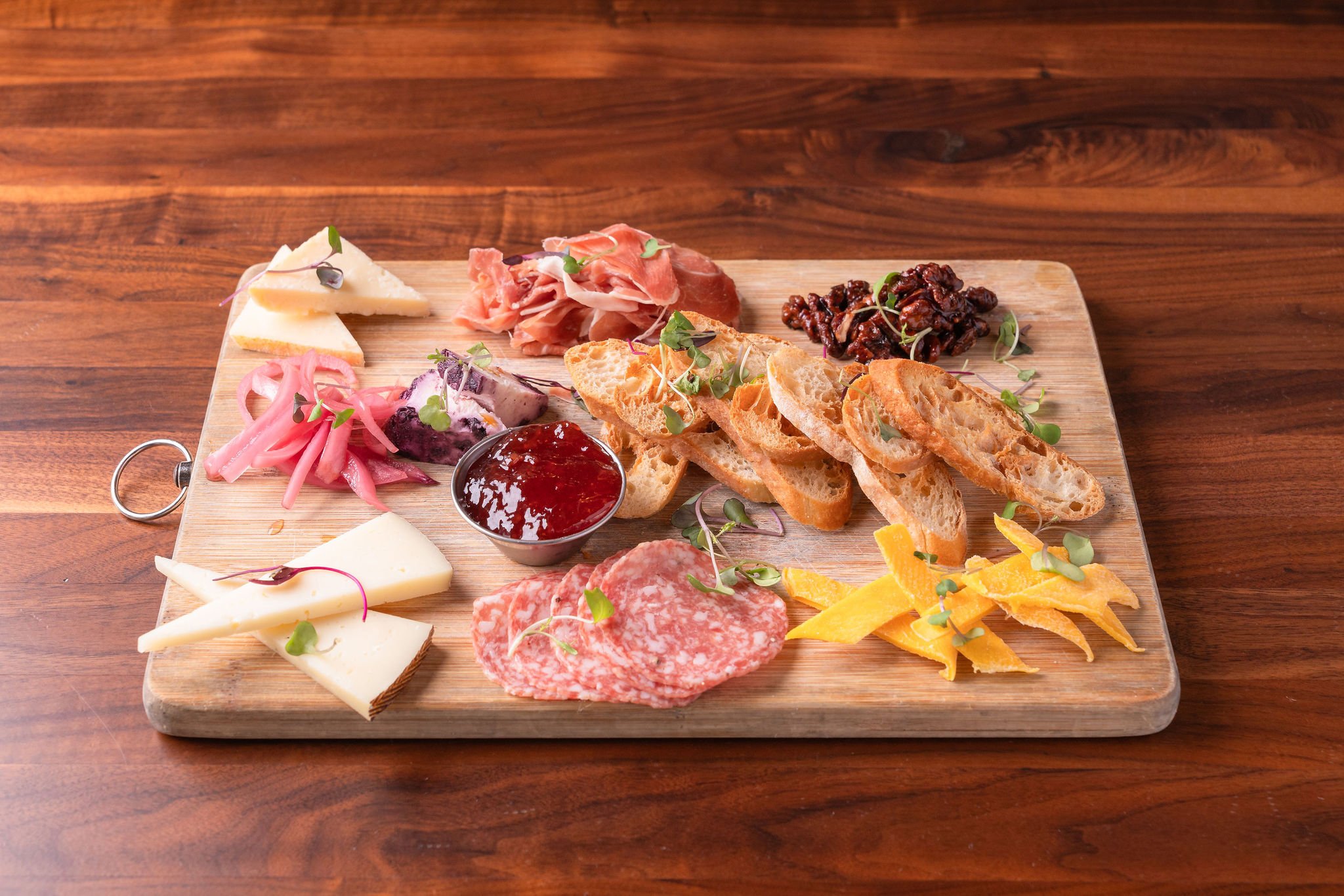 A wooden cheese (charcuterie) board with assorted cheeses, cured meats, slices of baguette, pickled vegetables, jam, and microgreens.